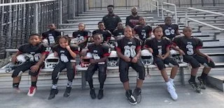 A youth football team sitting on bleachers, wearing black and white uniforms, with some players holding helmets and footballs.