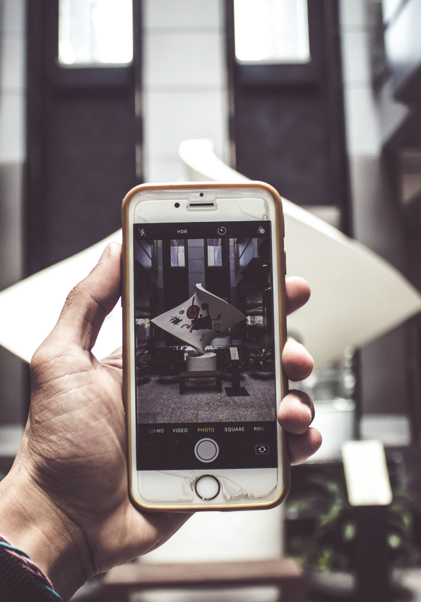 Person holding a smartphone capturing a photo of a contemporary art sculpture with a diamond-shaped base and abstract design, in a modern interior space with large windows.
