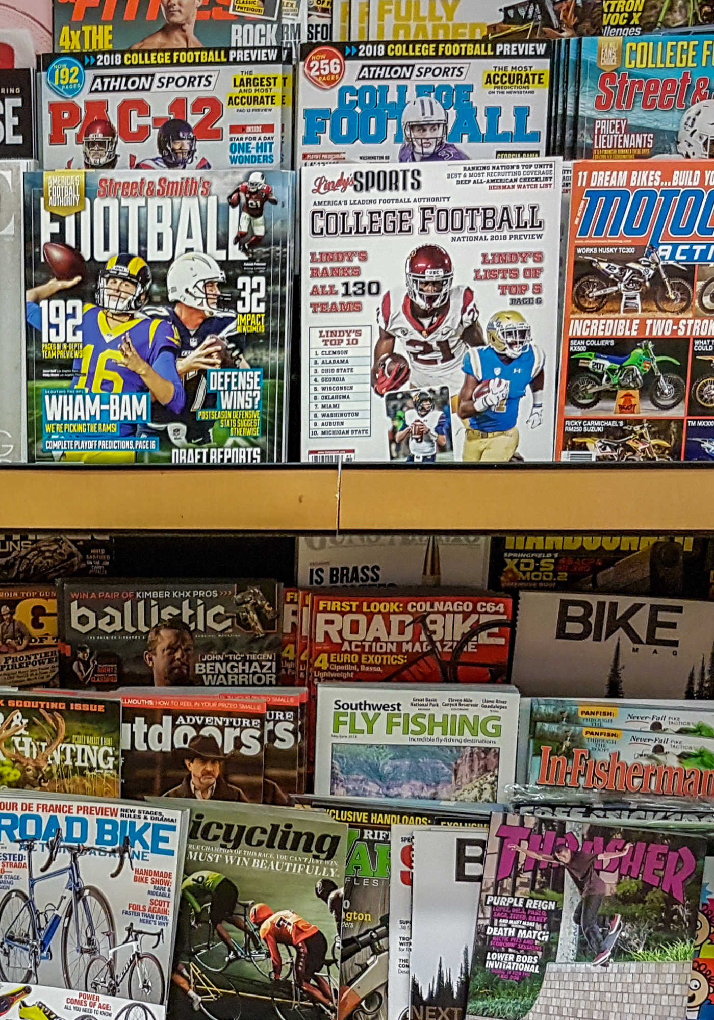 A display of sports and outdoor magazines on a rack, featuring football magazines with players, a motorcycle magazine, a cycling magazine, and fly fishing magazine, among others.