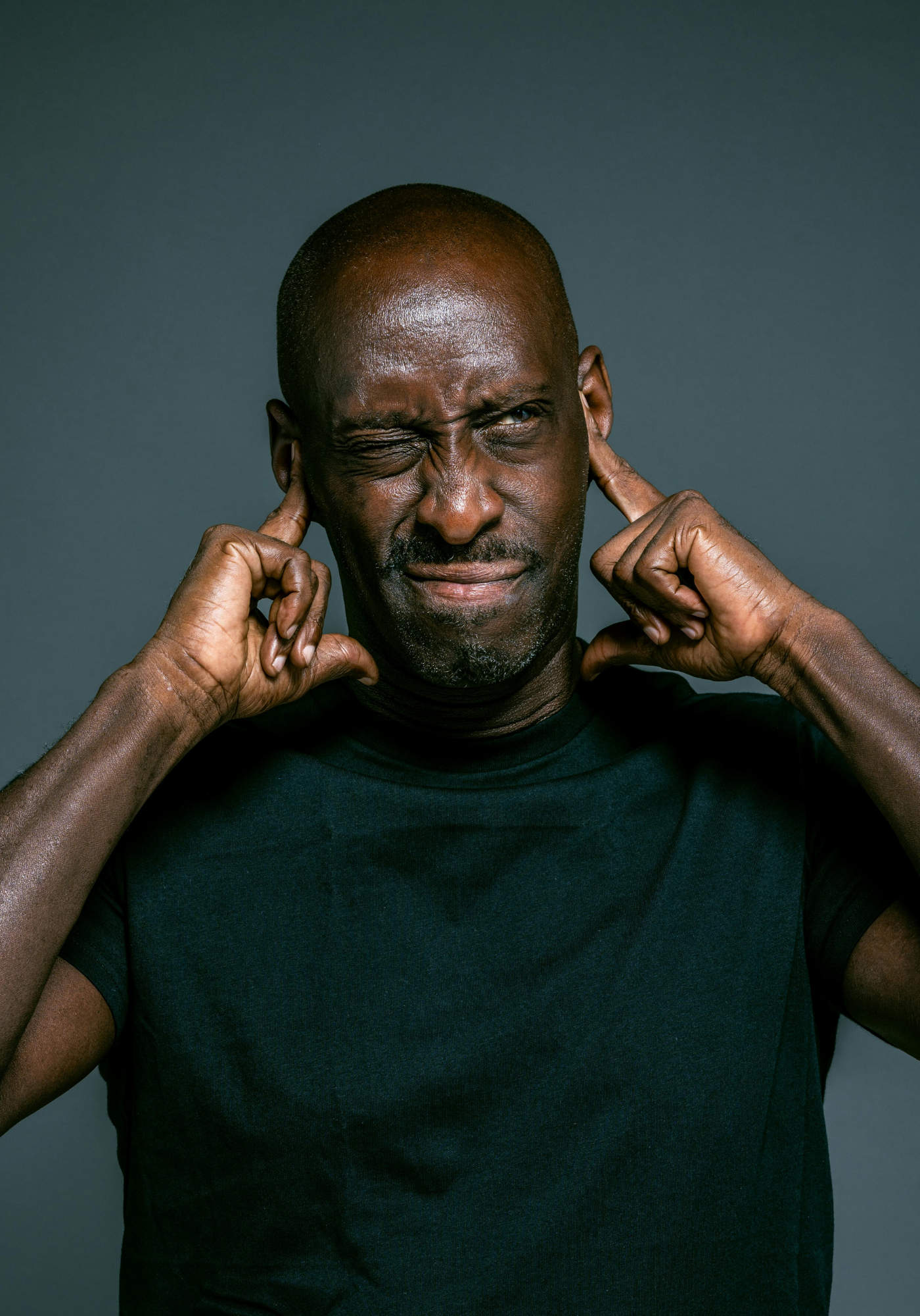 A man with a shaved head and dark skin covering his ears with his fingers, squinting one eye and making a pained expression against a plain gray background.