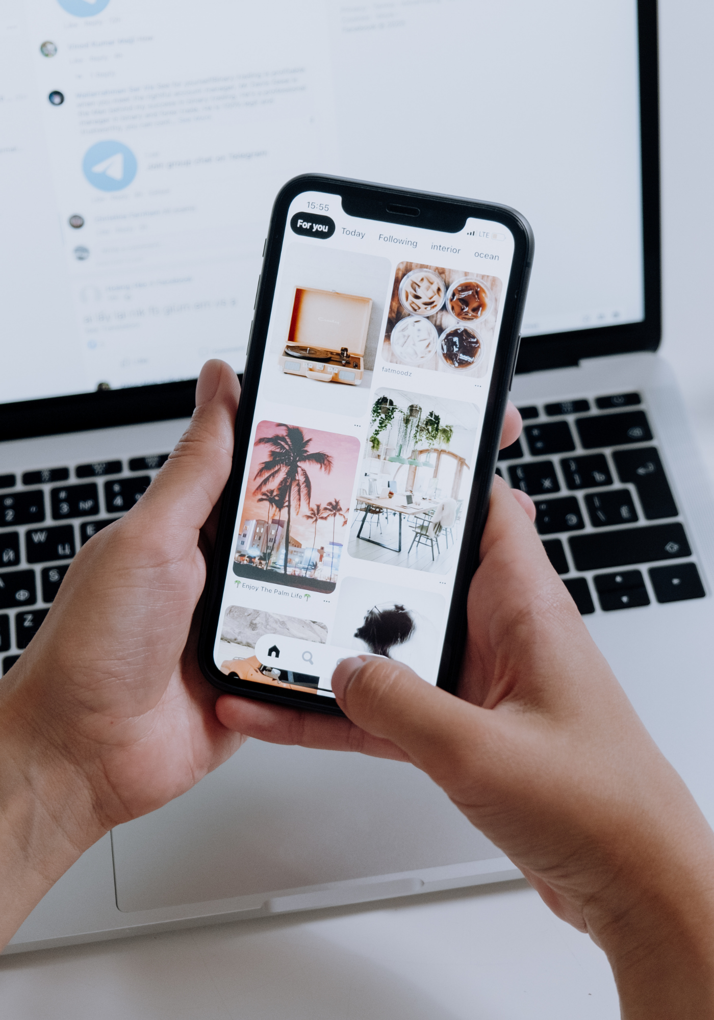 Person holding a smartphone displaying a Pinterest app with images of drinks, palm trees, and a workspace, in front of a laptop on a desk.