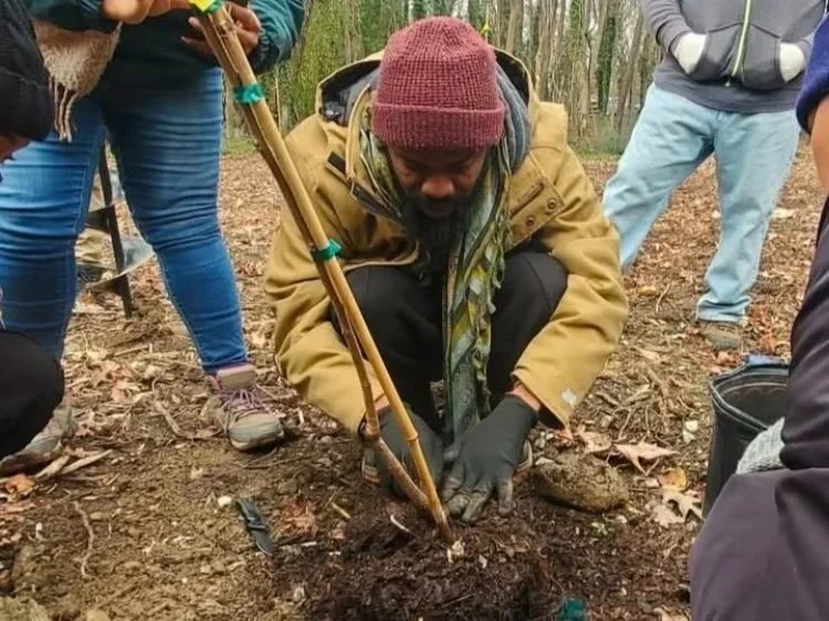 Duron Chavis, founder of Happily Natural, kneels to plant a sapling in the ground while a group of volunteers surrounds him.