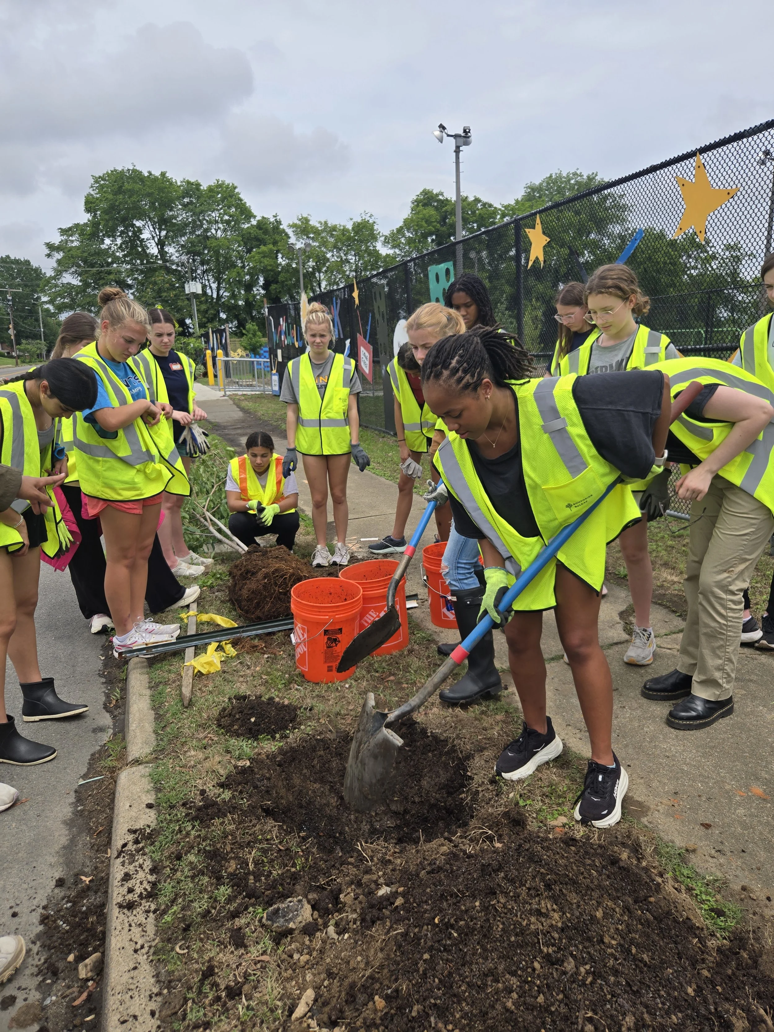 Two students demonstrate how to incorporate biochar into the soil using shovels while their peers watch and learn.