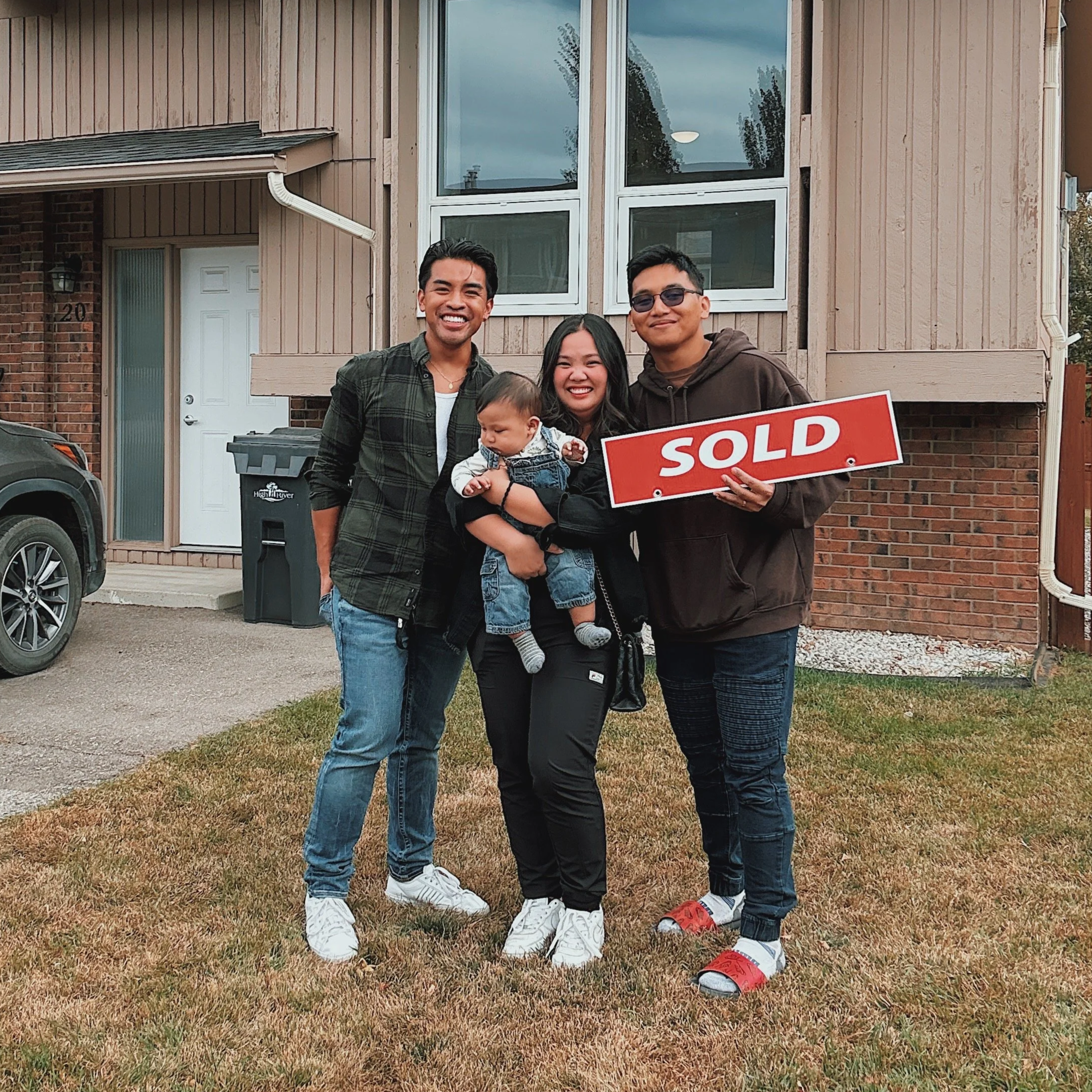 A group of four people, including a child, standing in front of a house with brown and brick exterior, holding a "SOLD" sign, smiling.