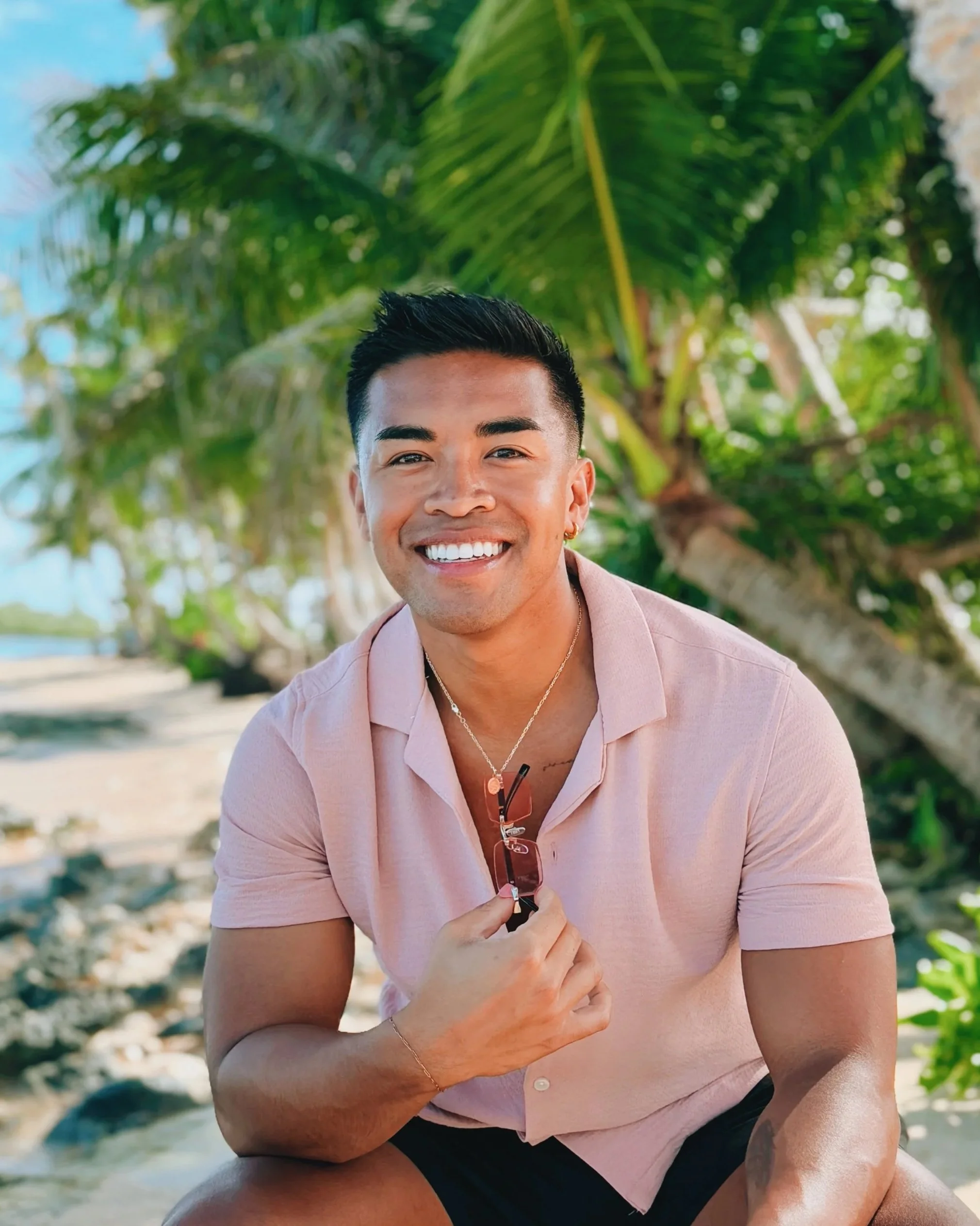 Smiling man in pink shirt holding sunglasses on a beach with palm trees.