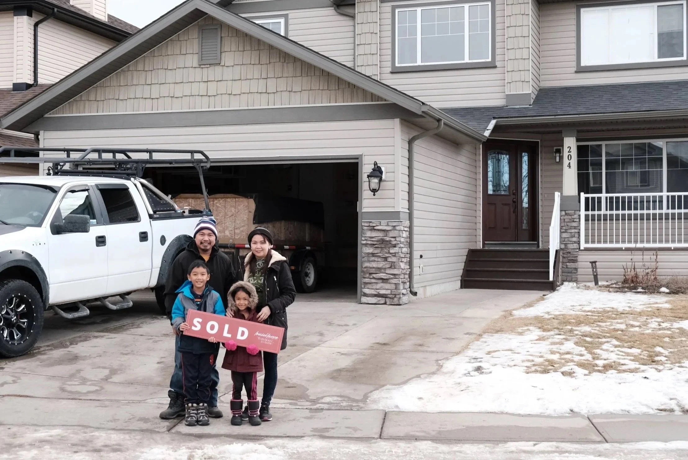 Family of four standing in front of a newly sold house, holding a 'SOLD' sign, with a white truck parked in the driveway, snow on the ground, and a beige house with stone accents.