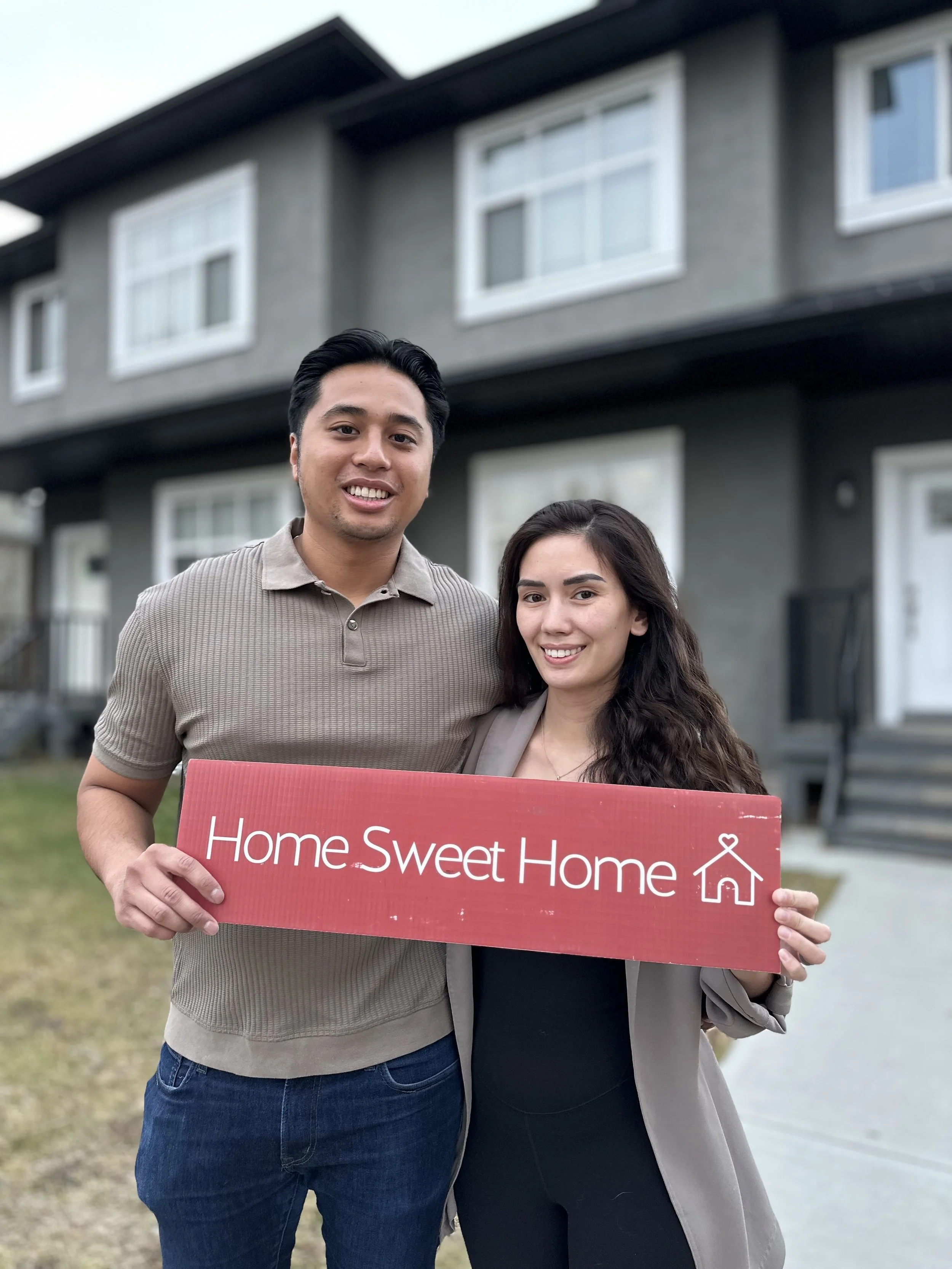 A happy couple standing in front of a house, holding a red sign that says 'Home Sweet Home'.