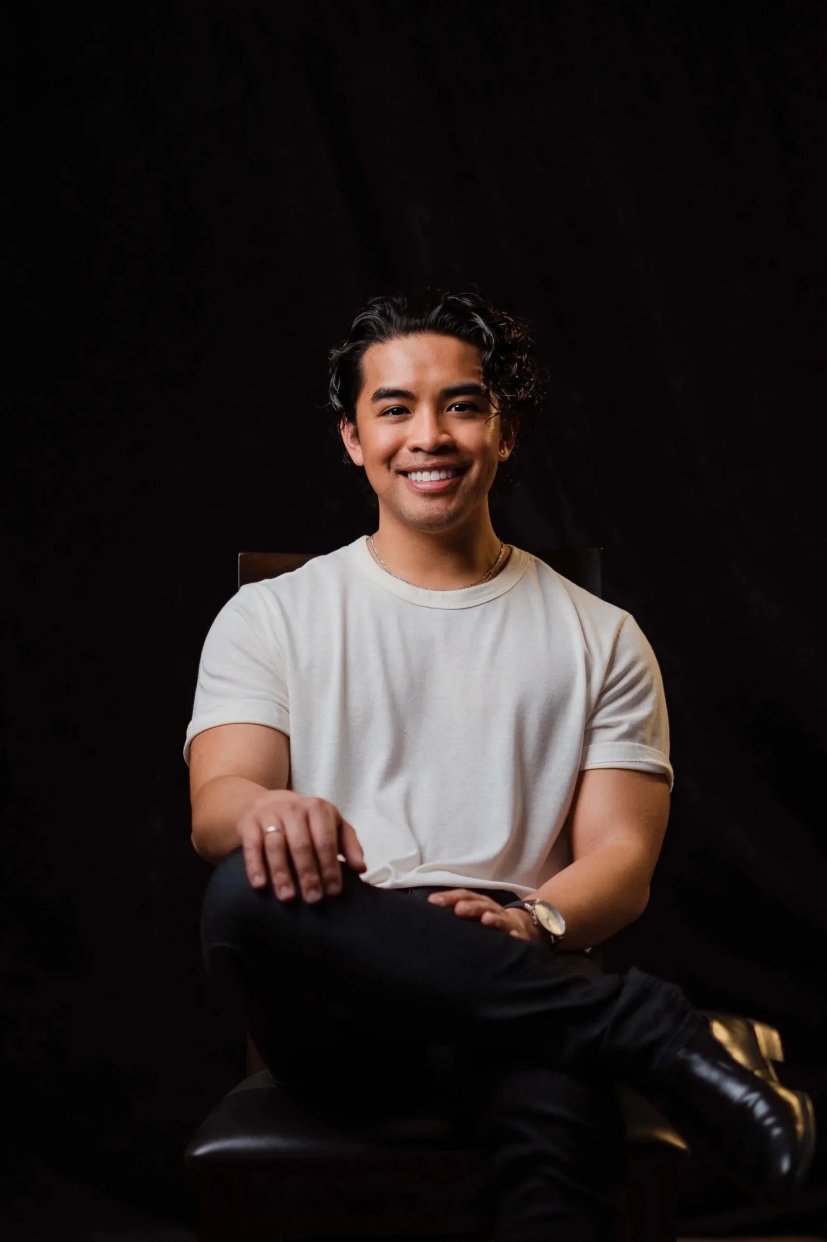 A young man with curly dark hair, wearing a white t-shirt and black pants, is seated on a chair against a black background, smiling at the camera.