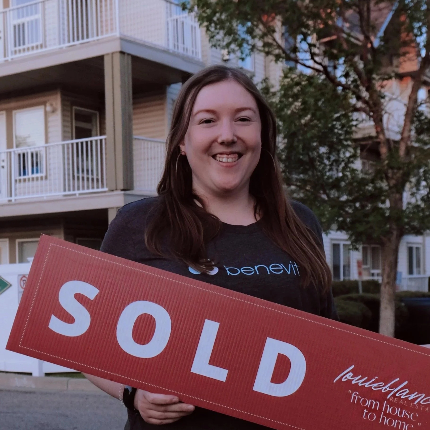 A woman with long brown hair holding a red 'SOLD' sign outside residential buildings with trees in the background.