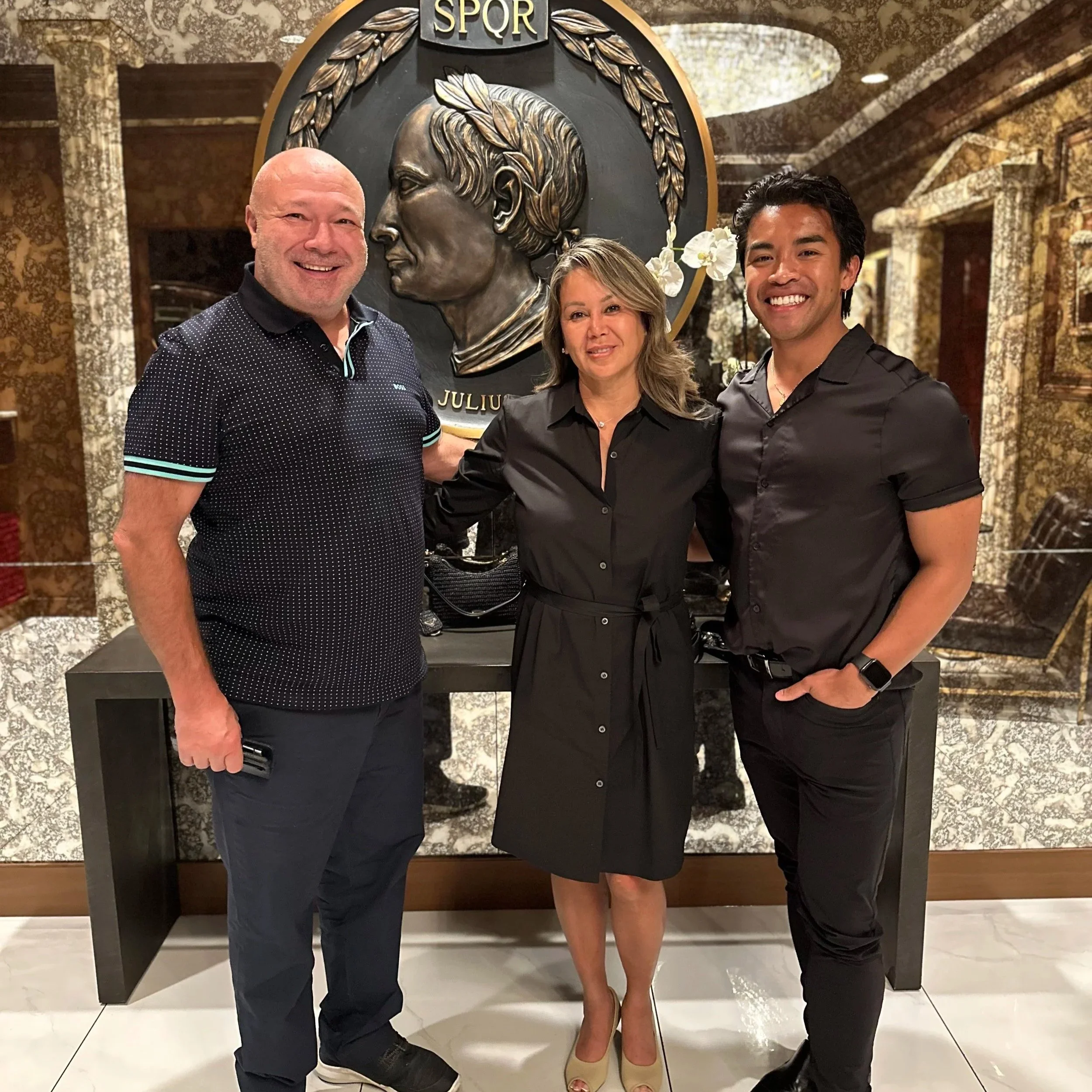 Three people standing together in front of a large bronze portrait of Julius Caesar with the acronym SPQR above it in a decorative setting.