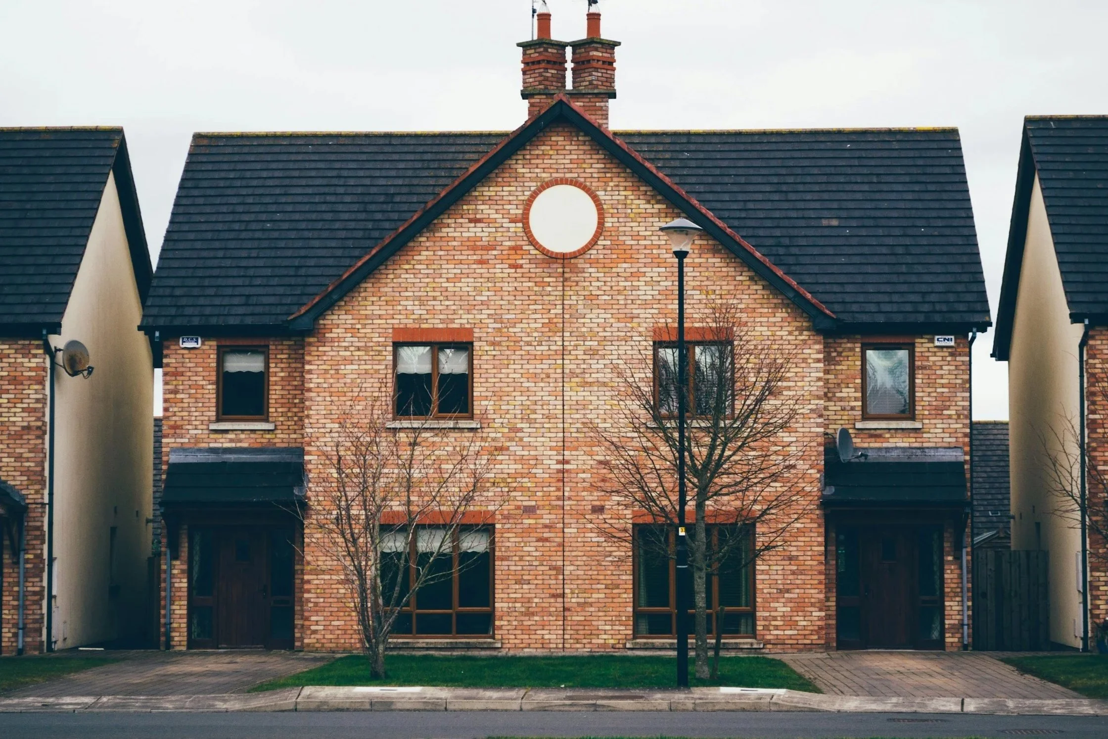A two-story brick residential building with a gabled roof, two chimneys, and three windows on each floor. There are two leafless trees and a lamppost in front, with driveways on either side leading to the back.