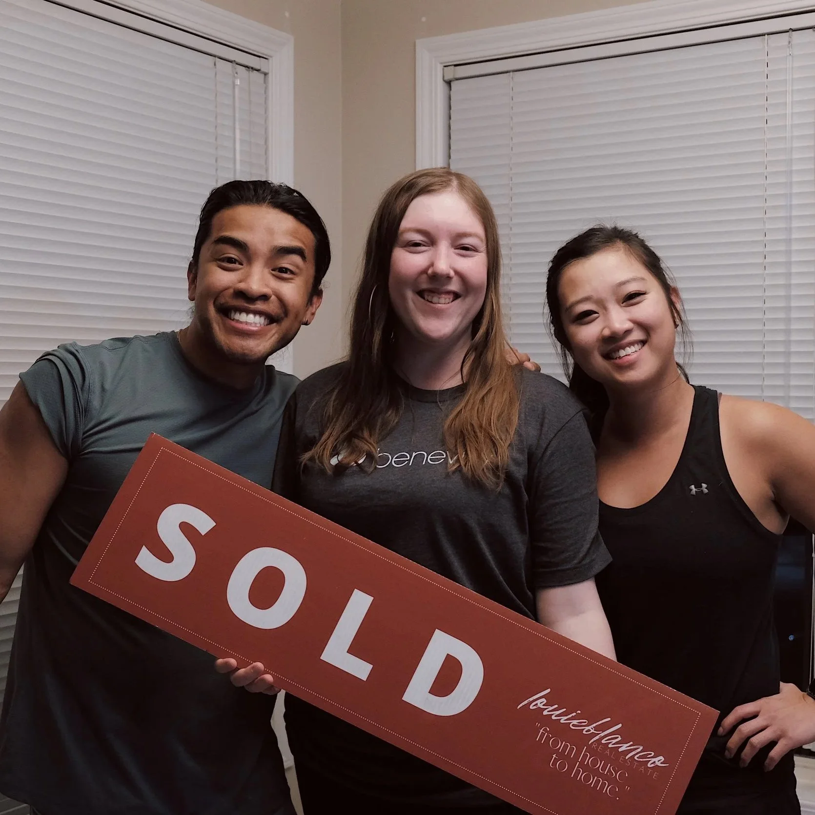 Three smiling friends holding a large red sign that says "SOLD" inside a room with closed white blinds on the windows.