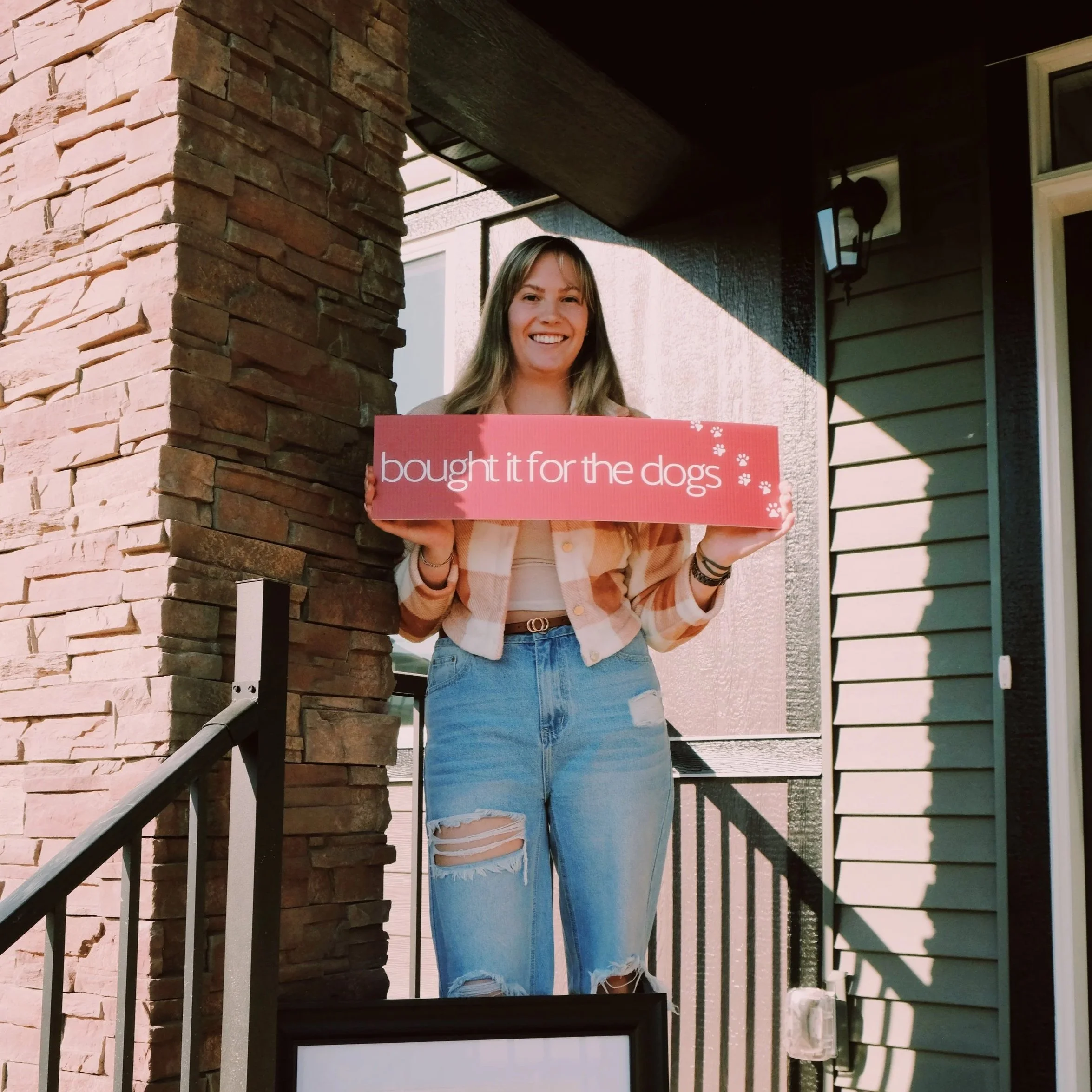 A young woman standing on a front porch holding a pink sign that says "bought it for the dogs" with a smile. She has long hair, wears a plaid jacket, a white top, and ripped jeans. The porch has a stone column and a dark-colored wall with a lamp above.