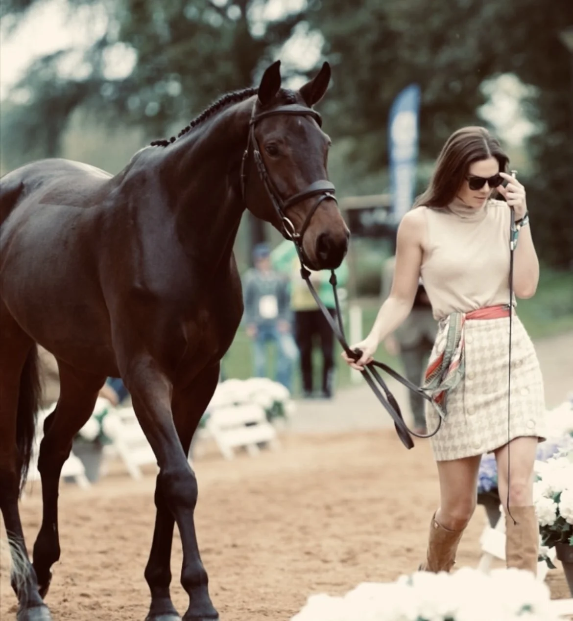A woman in sunglasses, a beige top, and a plaid skirt walking a dark brown horse on a leash outdoors during daytime.