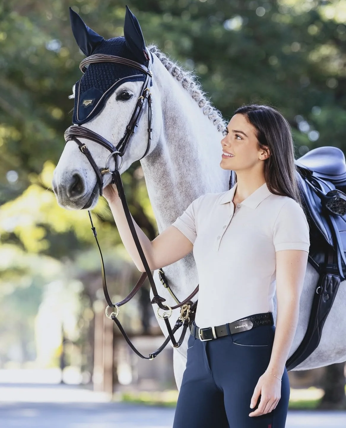 A woman petting a white horse outside on a sunny day, with trees in the background.