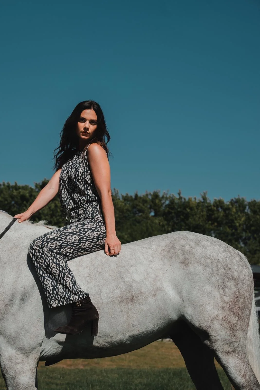 A woman with long dark hair wearing a black and white patterned jumpsuit and black high-heeled boots sitting on a white horse outdoors under a clear blue sky.