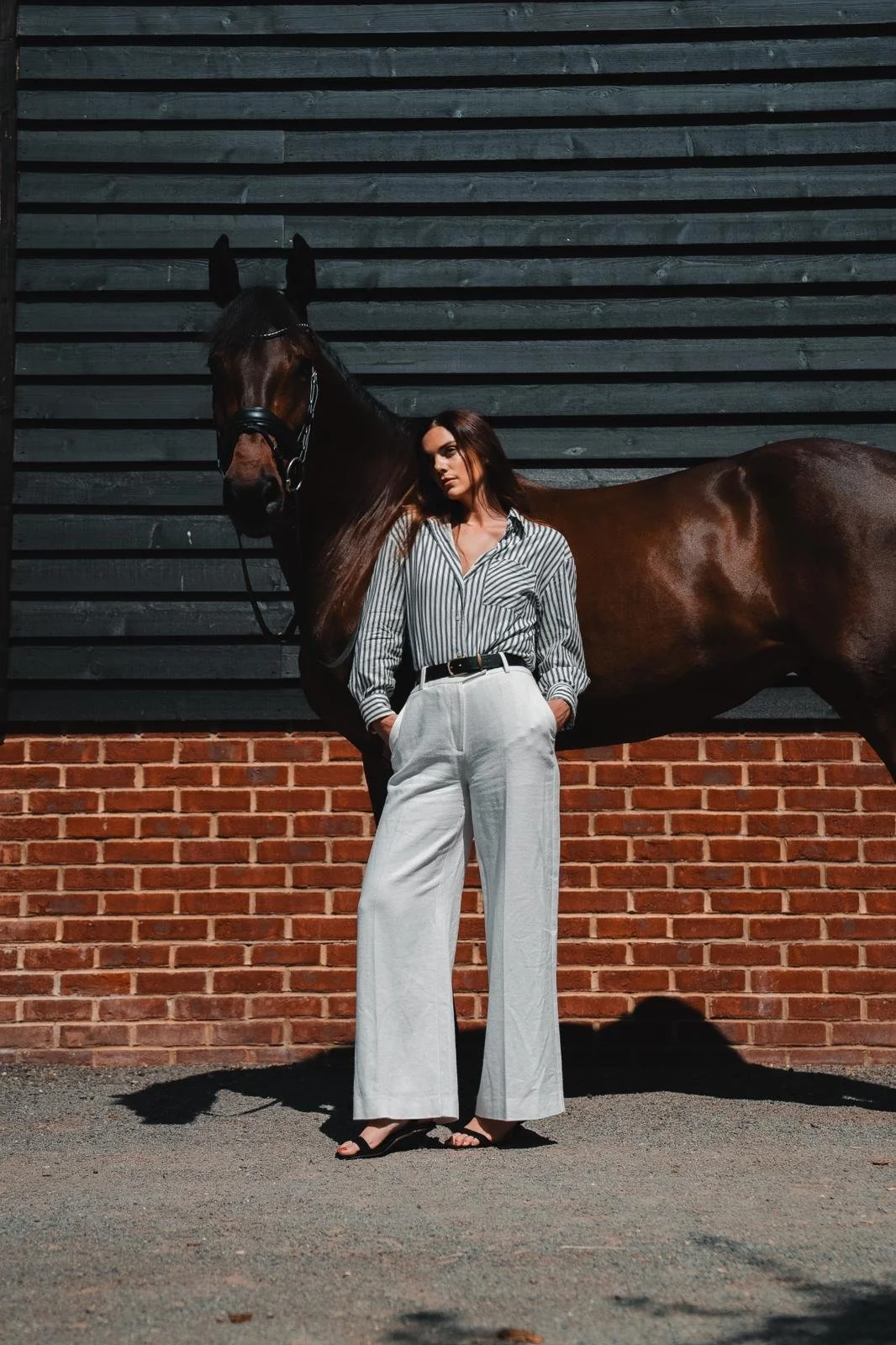 A woman standing next to a brown horse outdoors, with a black wooden wall and brick wall background, wearing a striped shirt, wide-leg white trousers, and sandals.