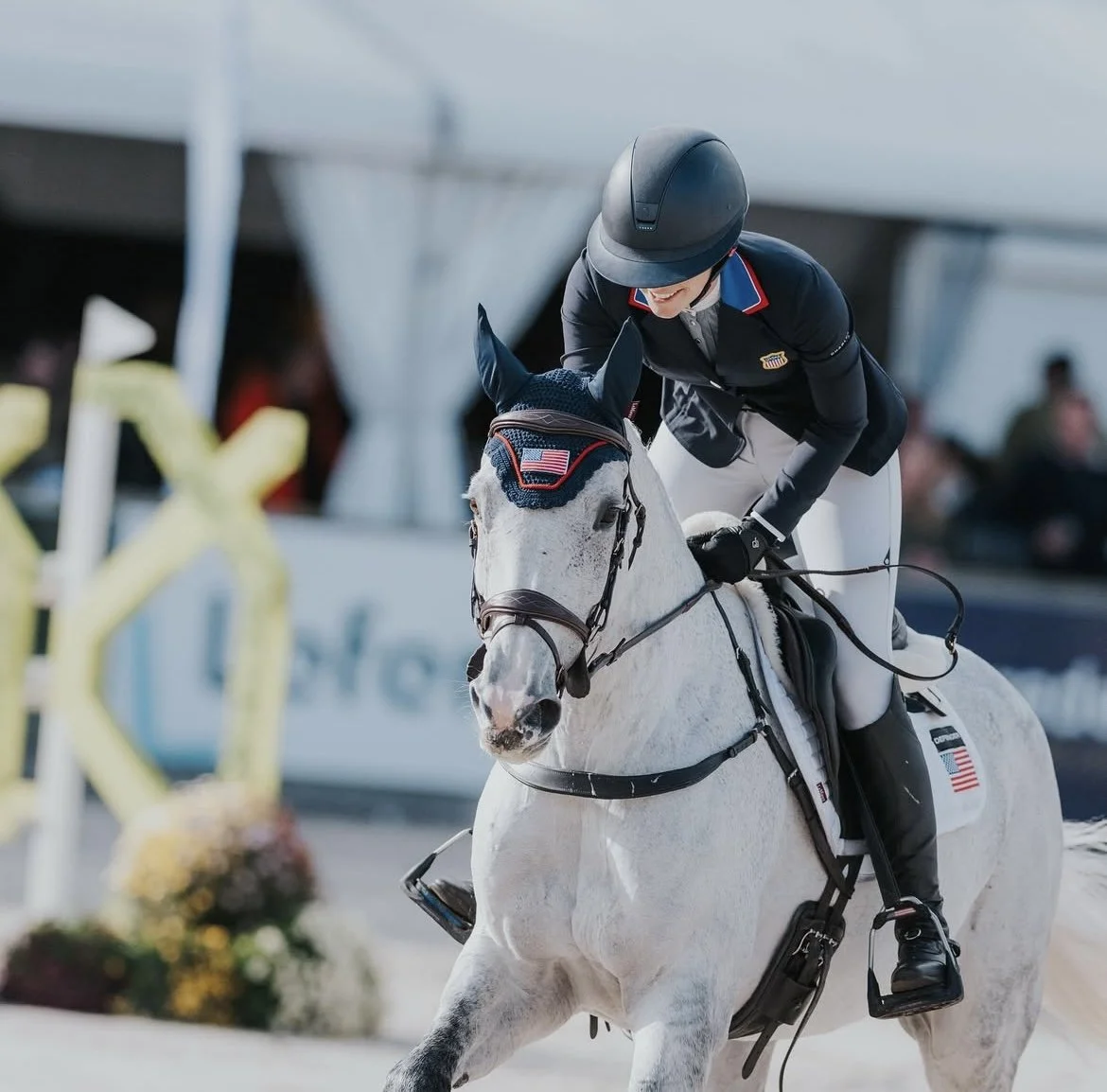 An equestrian athlete riding a white horse during a show jumping event, with a woman rider wearing a dark jacket, white breeches, and a helmet, smiling and holding the reins.