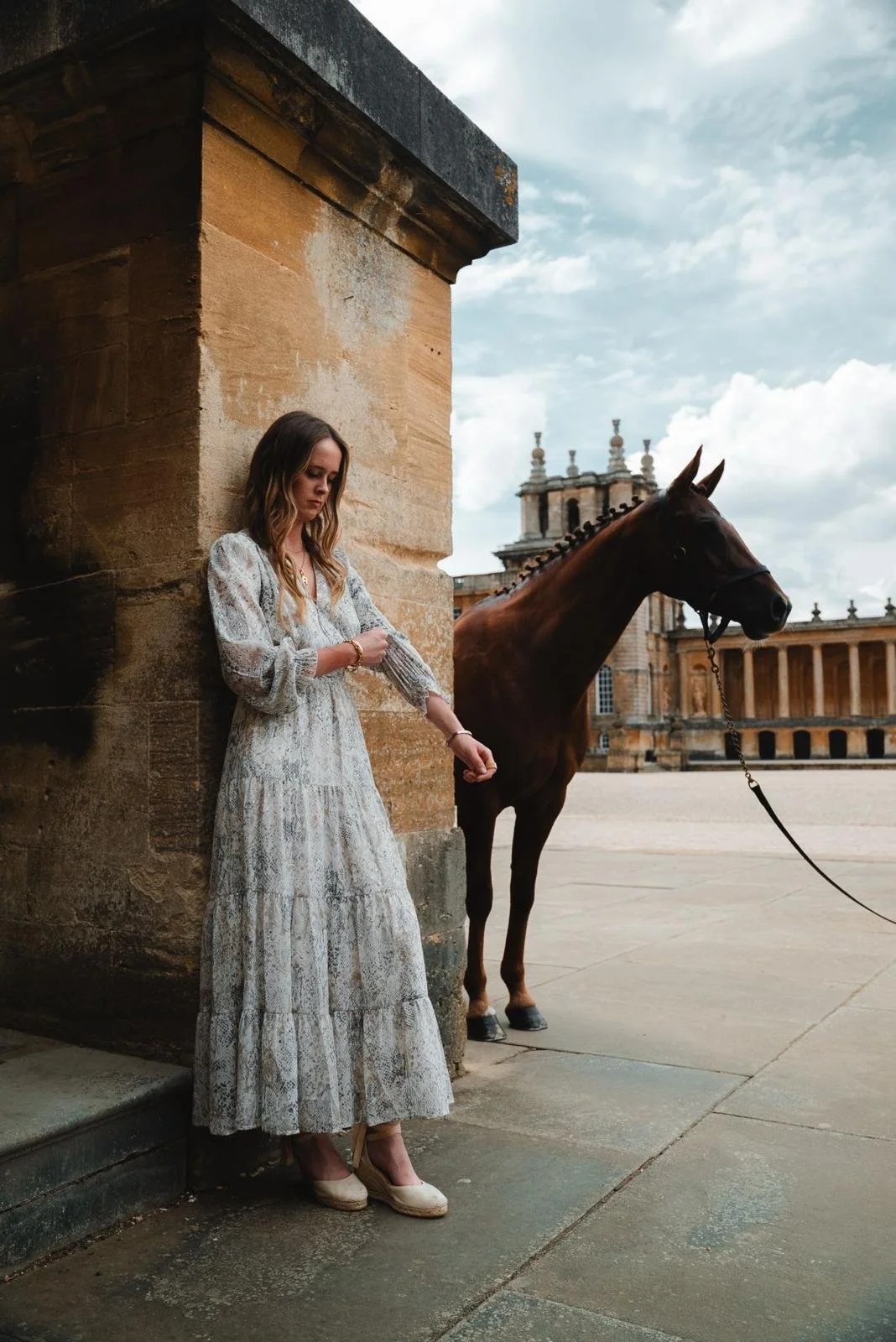 A woman in a long, light-colored lace dress and high heels stands against a stone wall, looking at her wrist, with a brown horse on a leash nearby in an open courtyard with historic buildings and a cloudy sky in the background.