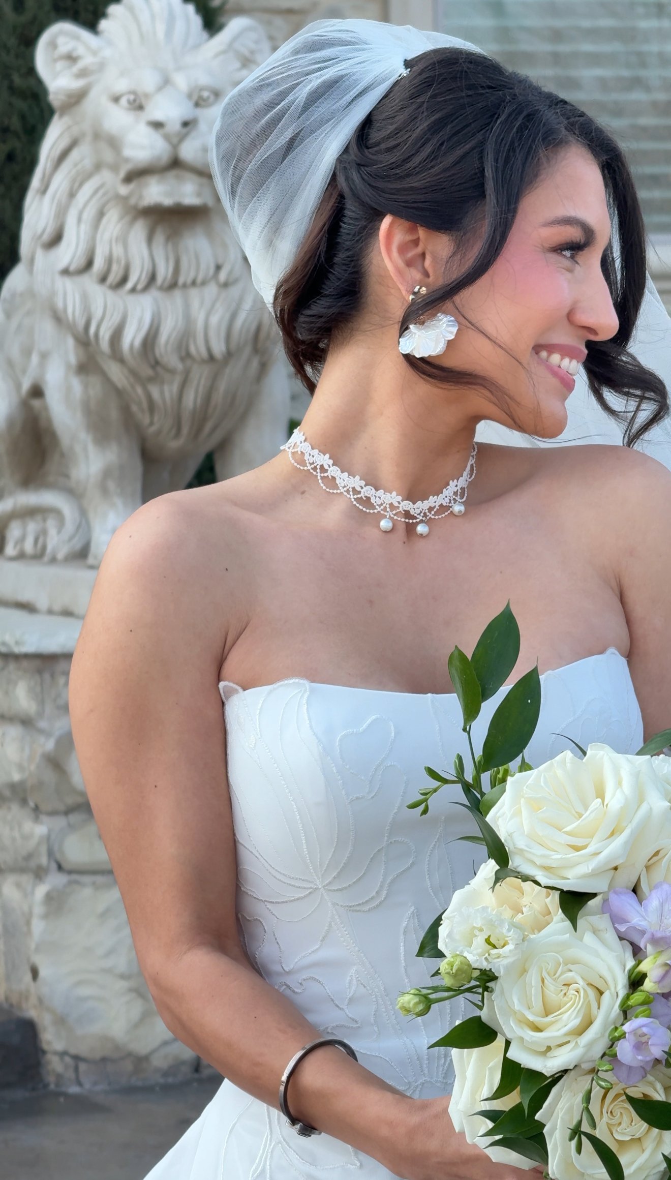 Outdoor bridal portrait of a bride in a strapless embroidered wedding gown with pearl choker necklace and veil, holding a white rose bouquet for a romantic wedding day photo.