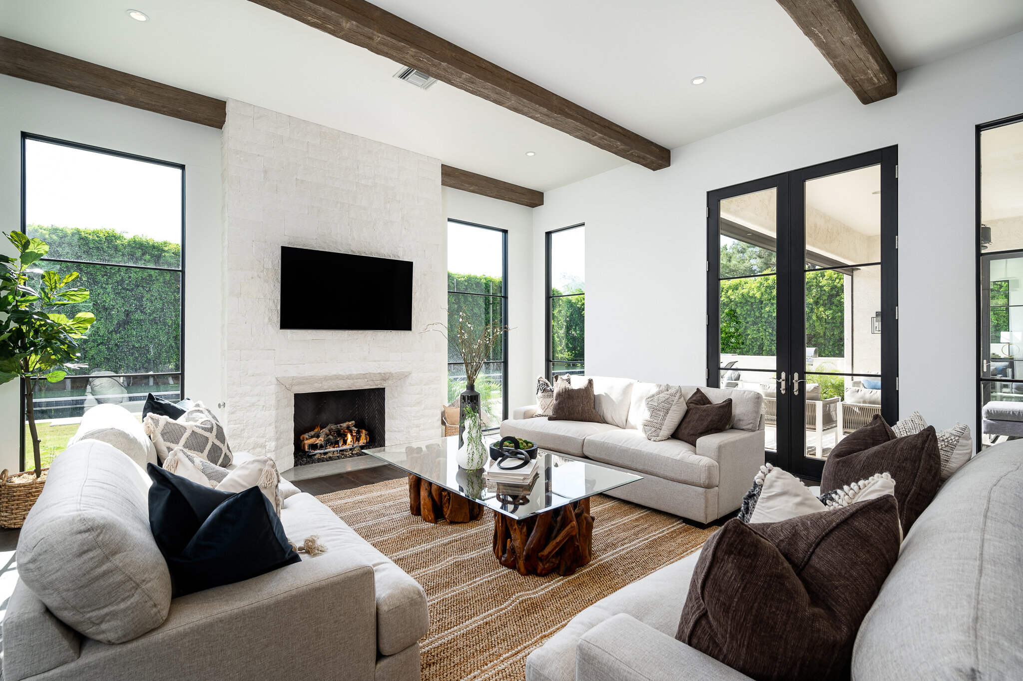 Modern living room with white sofas, a glass coffee table, a fireplace, large windows, and a flat-screen TV above the fireplace.