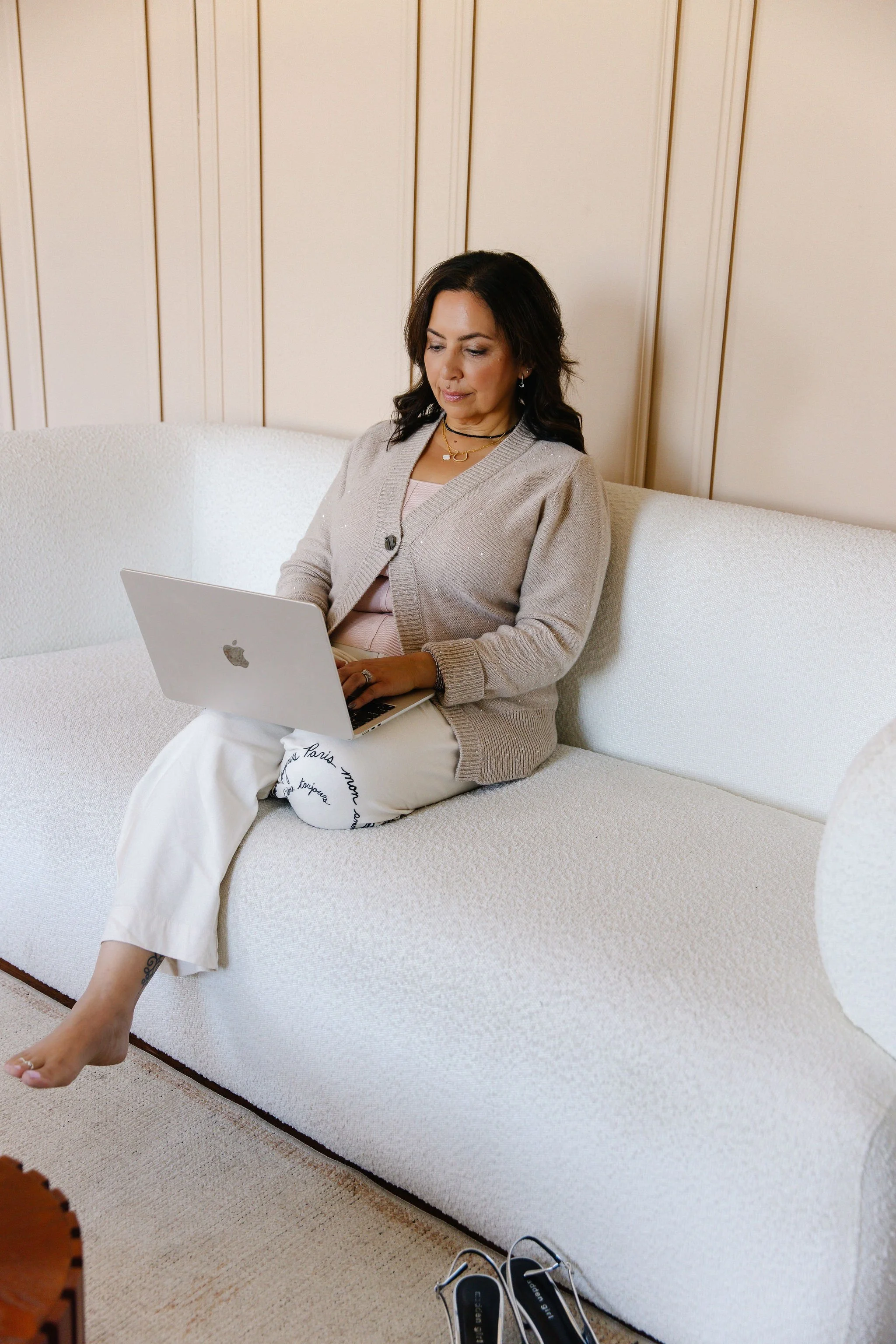 A woman sitting on a white sofa, working on a silver MacBook in a room with wooden wall paneling. She is wearing a beige cardigan, white pants, and has multiple necklaces. Her shoes are on the floor next to the sofa.