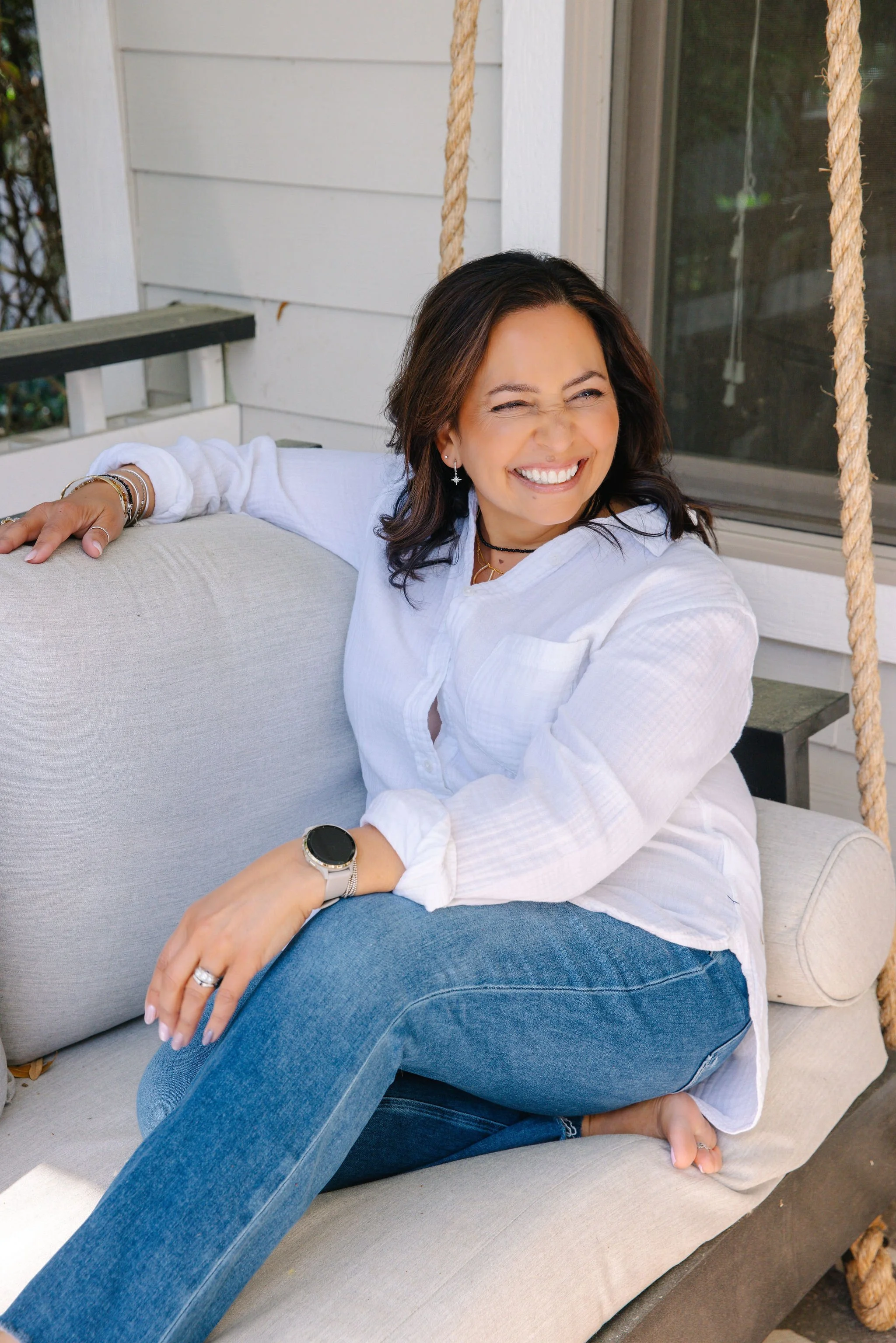 A woman with dark hair, wearing a white shirt and blue jeans, is sitting on a beige outdoor sofa, smiling and squinting in sunlight on a porch with white siding and a window with a screen.