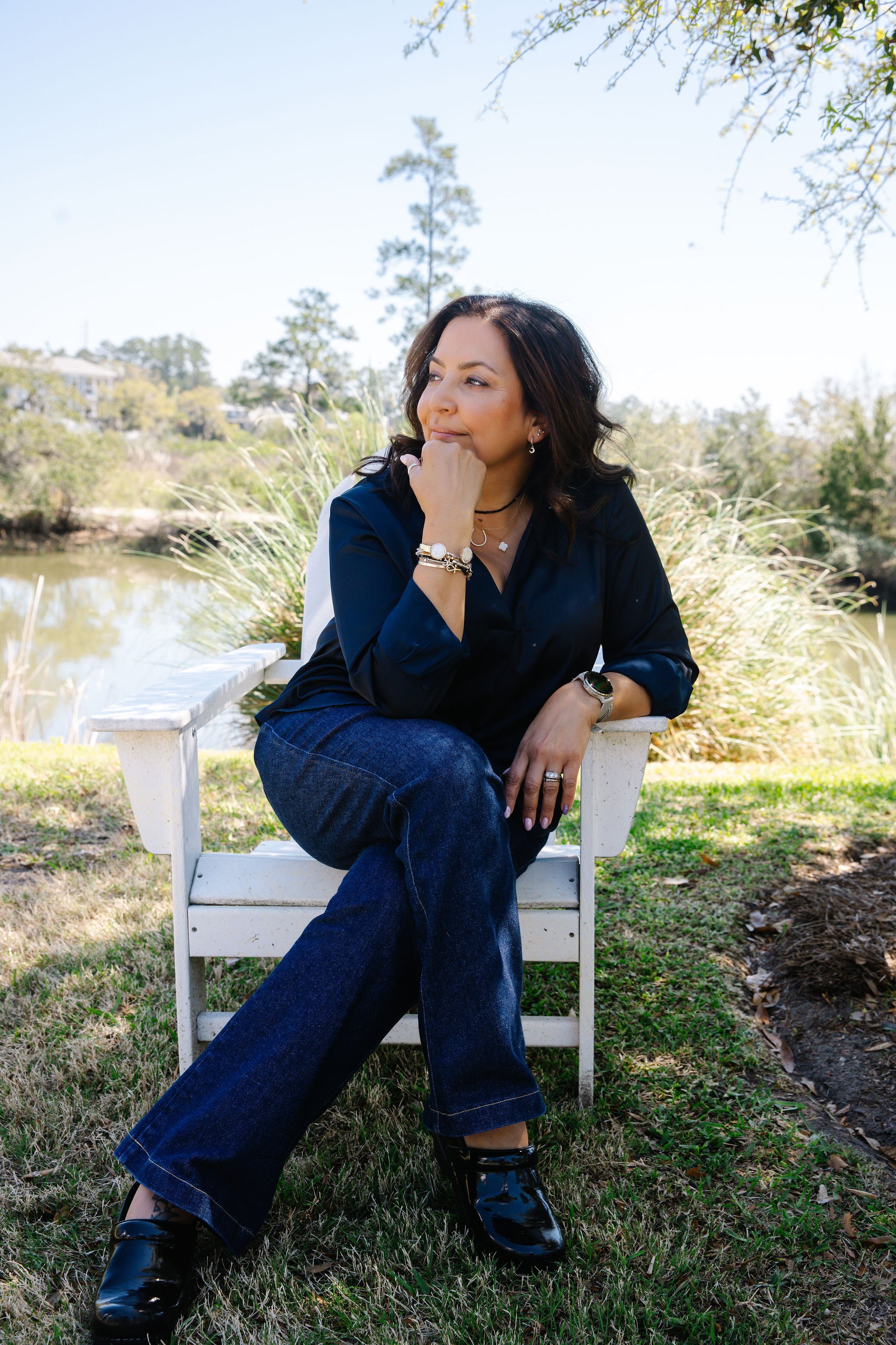 A woman with dark hair, wearing a dark blue blouse, jeans, and shiny black boots, is sitting on a white bench outdoors near a pond. She rests her chin on her hand, looking thoughtfully to the side, with trees and a clear blue sky in the background.