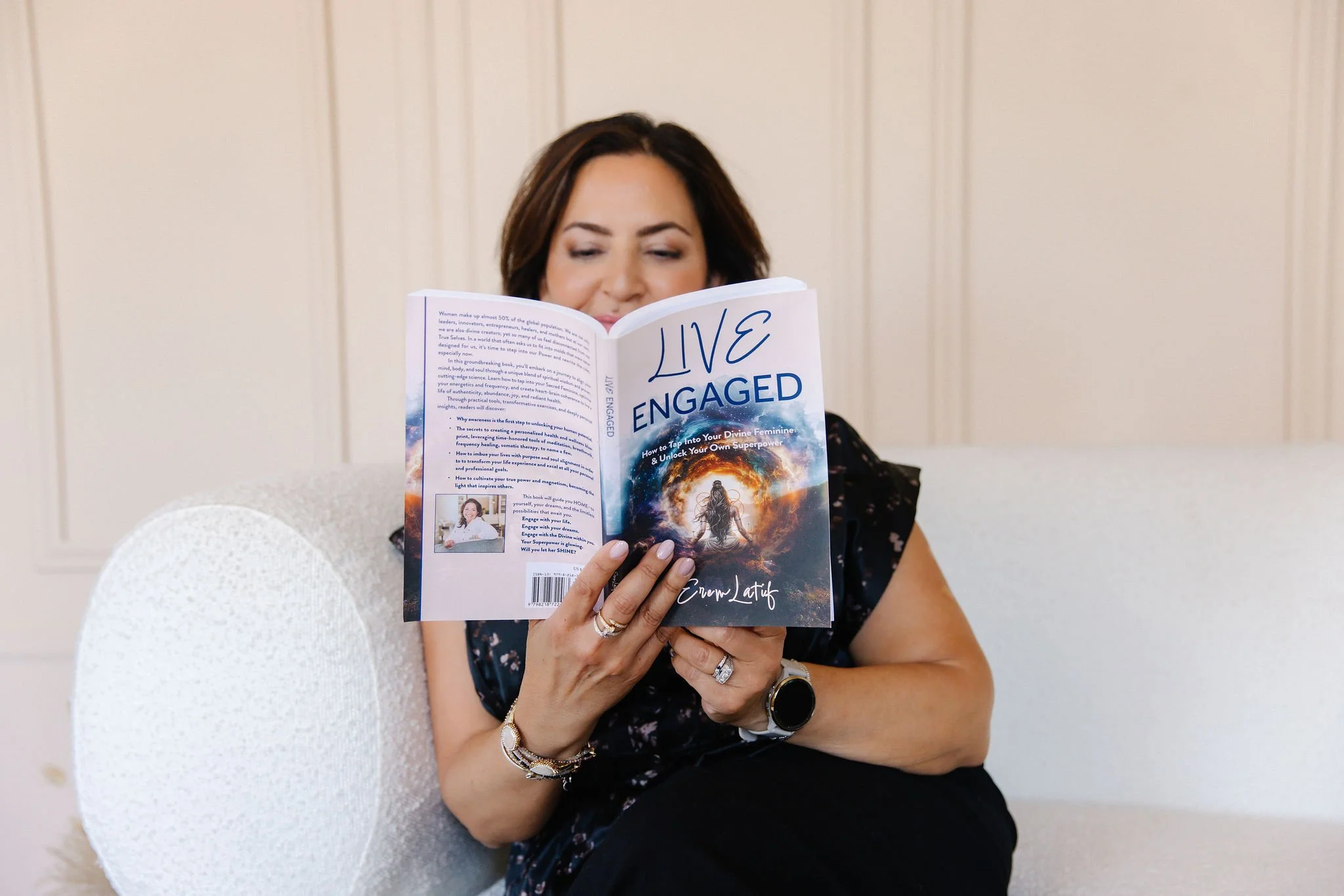 Woman with dark hair sitting on white chair, reading a book titled 'LIVE ENGAGED' with a cosmic scene on the cover, in a light-colored room.