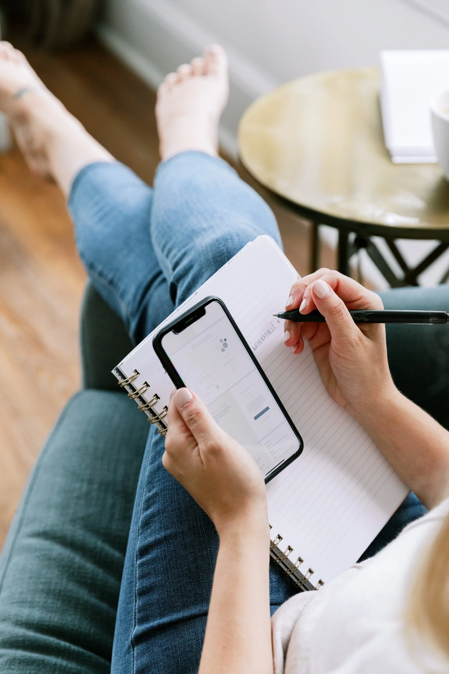 Person sitting on a green couch, holding a smartphone and taking notes in a notebook, with another person sitting next to them, visible only by their arm.