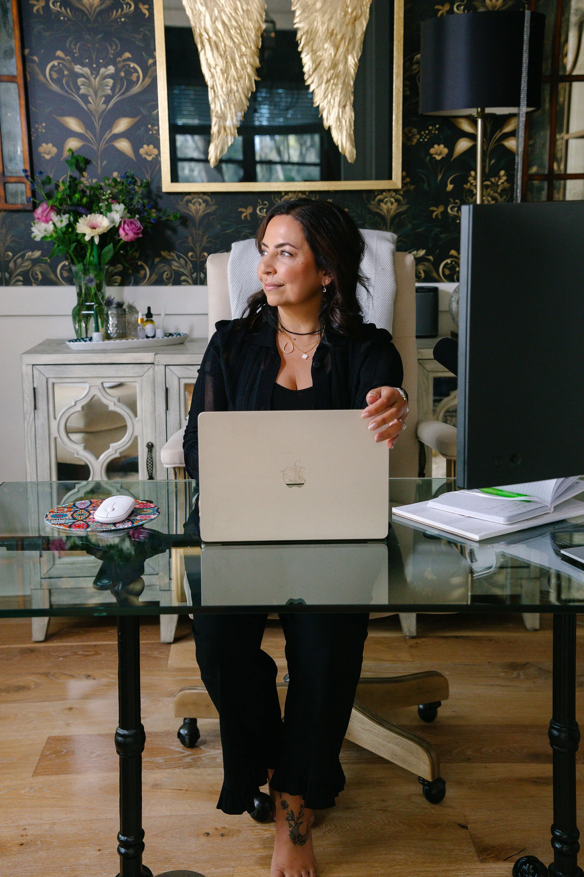 A woman sitting at a desk working on a laptop in a home office. She has dark hair, tattoos on her ankle, and is wearing a black outfit. There is a computer monitor, notebooks, and a mouse on the desk. Behind her, there is a decorative wall with a vase of flowers, a mirror with angel wings, and a black and gold wallpaper.
