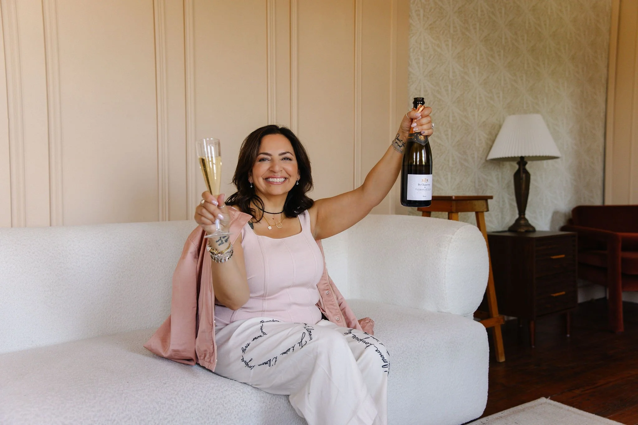 A woman with dark hair smiling and sitting on a white couch, holding a glass of champagne in one hand and a bottle of champagne in the other, in a living room with beige and patterned wallpaper, a lamp, and wooden furniture.