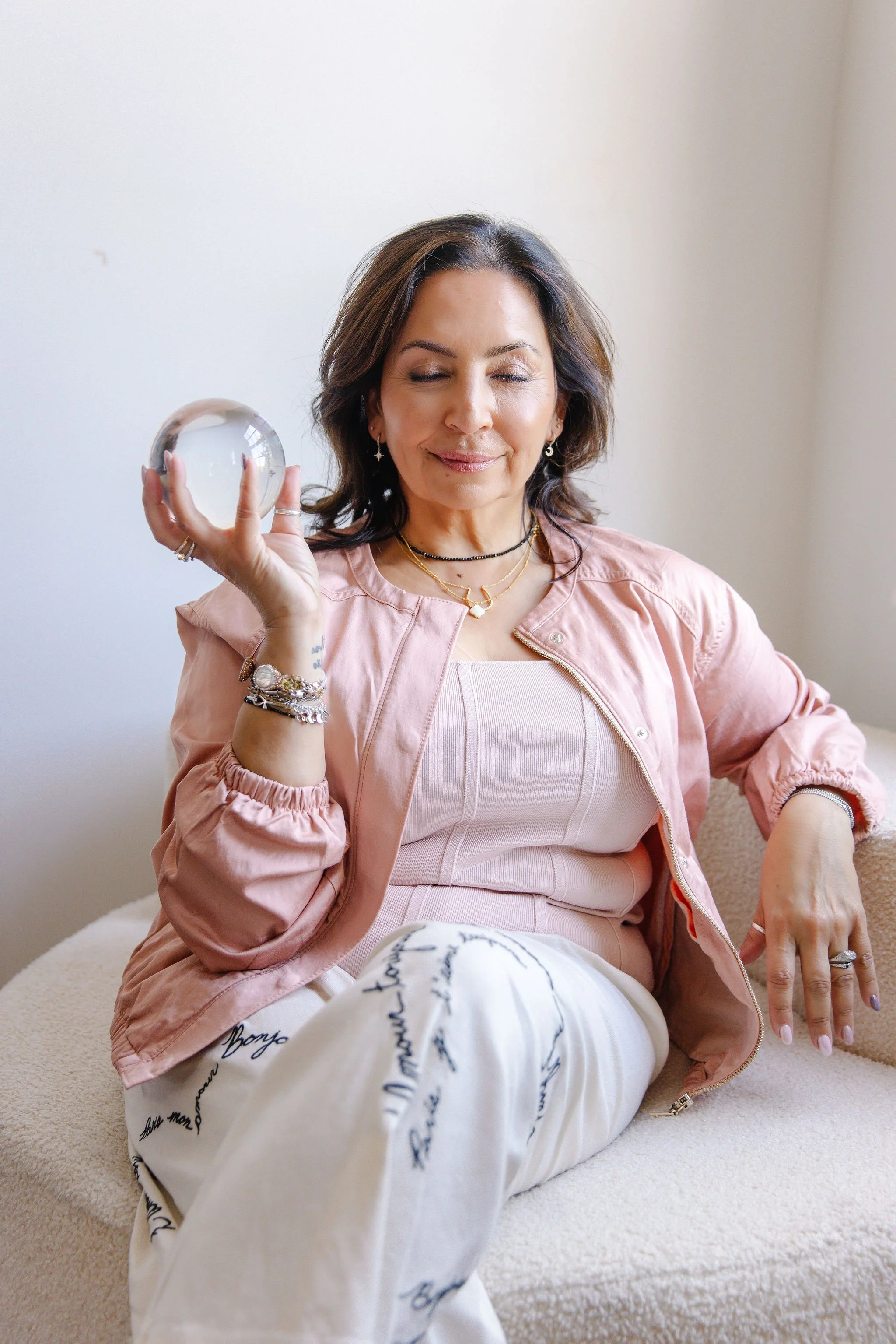 A woman with dark hair, wearing a pink jacket and layered necklaces, sitting with eyes closed, holding a glass orb in her right hand, and smiling softly.