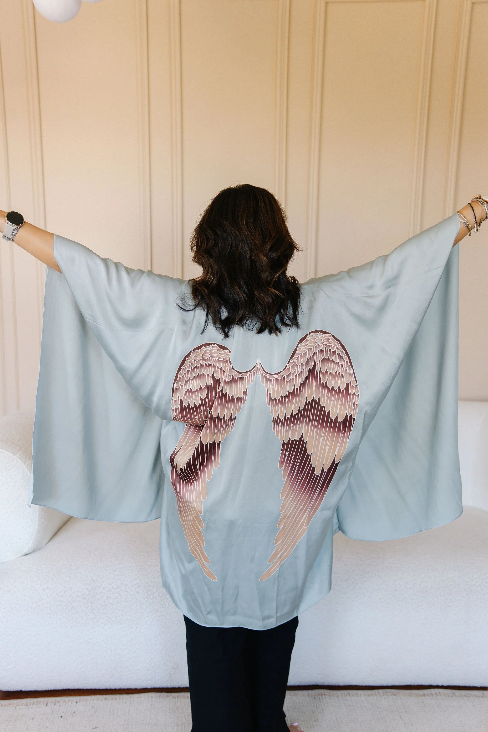 Person with medium-length curly dark hair and light skin wearing a loose, light blue or teal cape with a pair of angel wings printed on the back, standing indoors against a beige paneled wall.