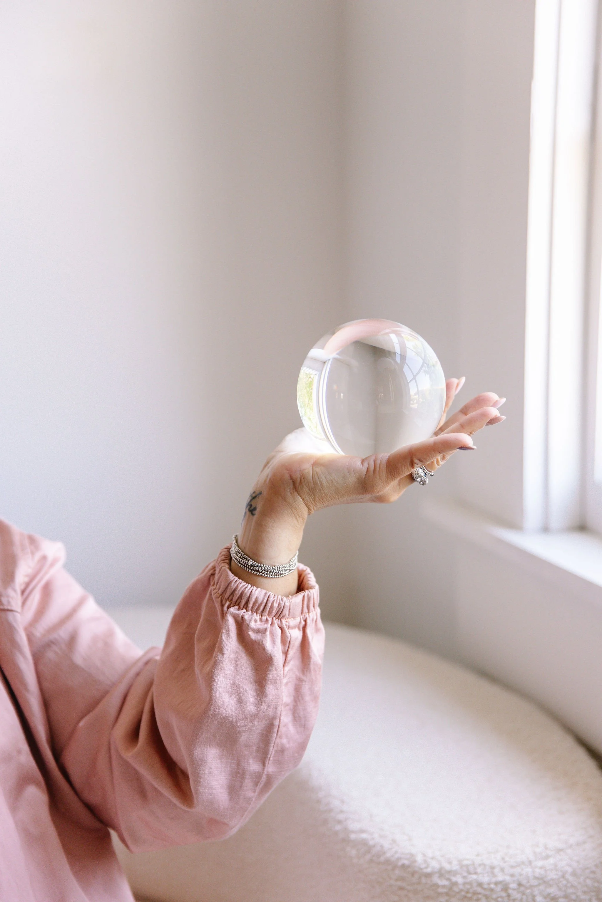 A person holding a clear glass sphere in their hand, wearing a pink long-sleeve shirt, with a beige wall and window in the background.
