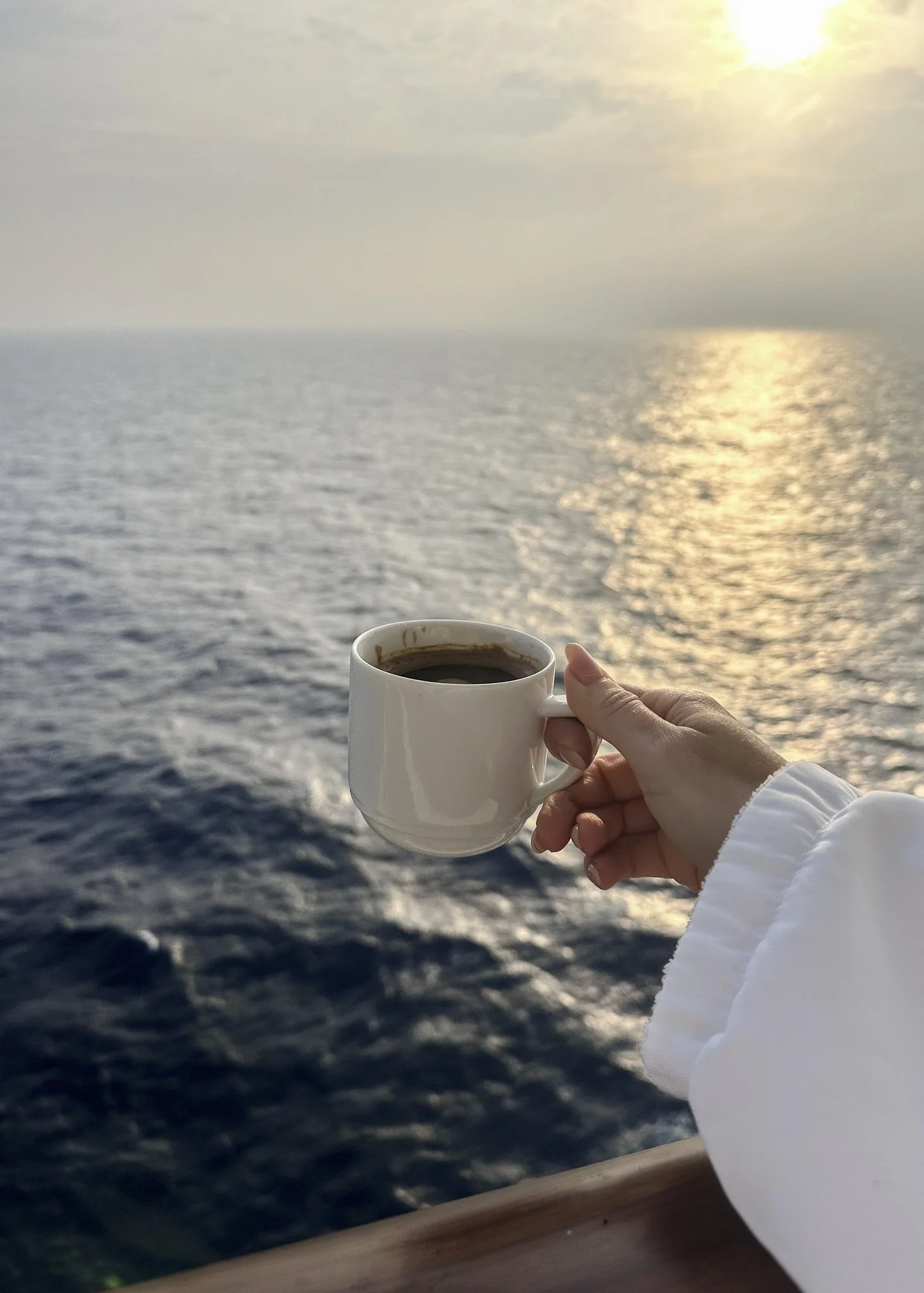 Person holding a white mug of coffee with an ocean and sunset in the background.