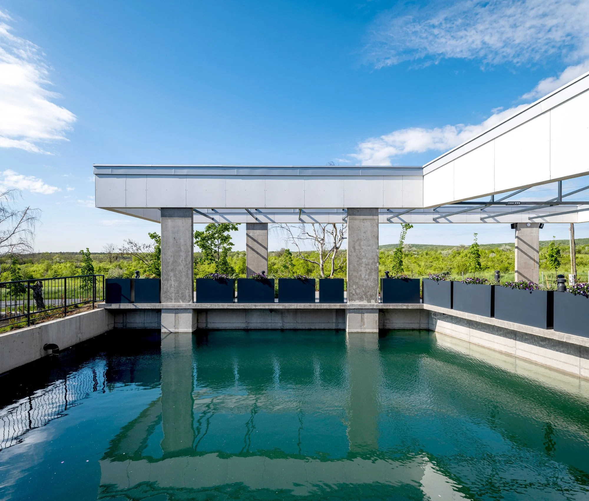Modern building with a glass roof over a water feature, surrounded by planters and a railing, with trees and a blue sky in the background.