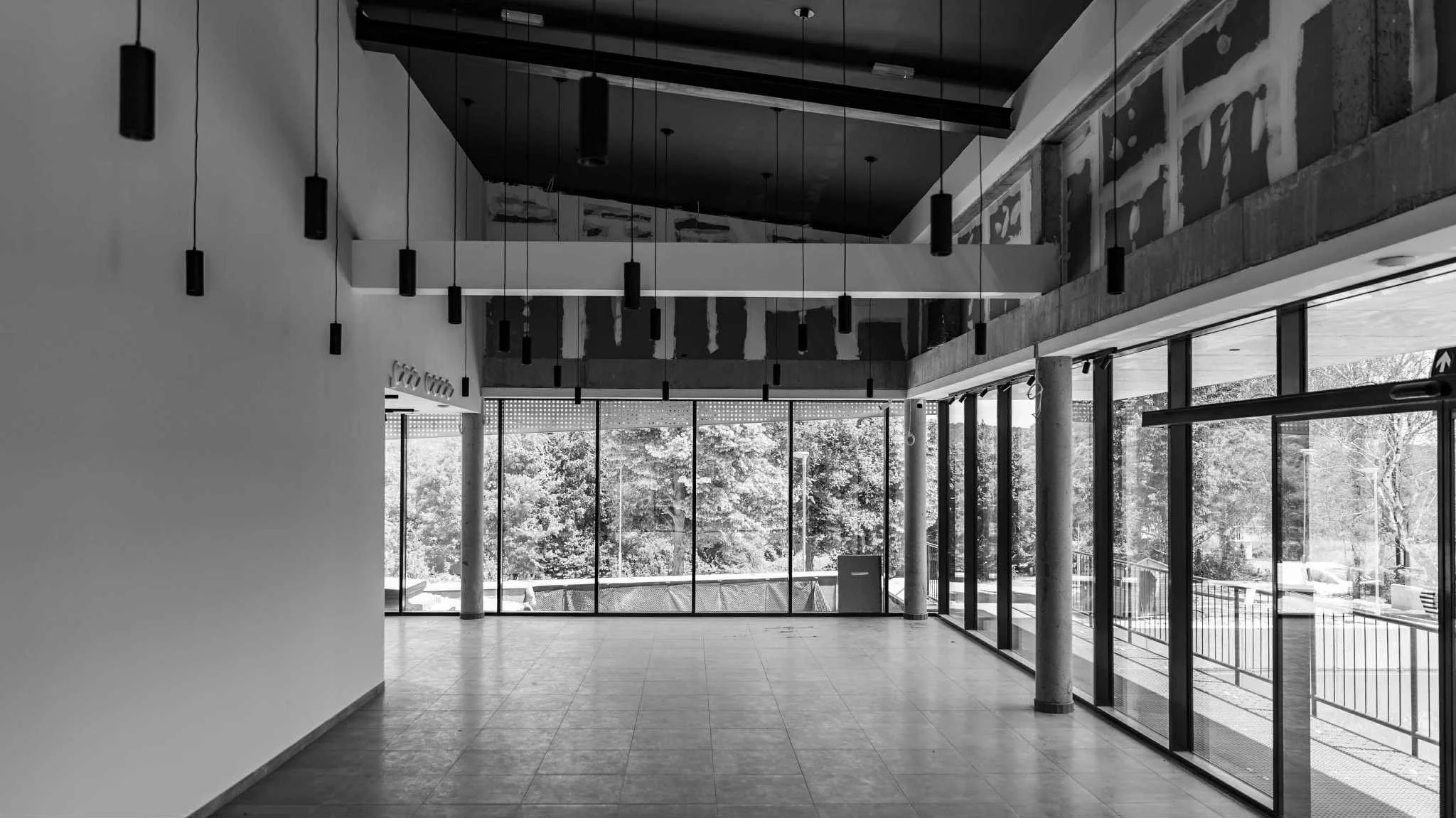 Empty modern interior space with large floor-to-ceiling windows, hanging pendant lights, and trees visible outside in daylight.