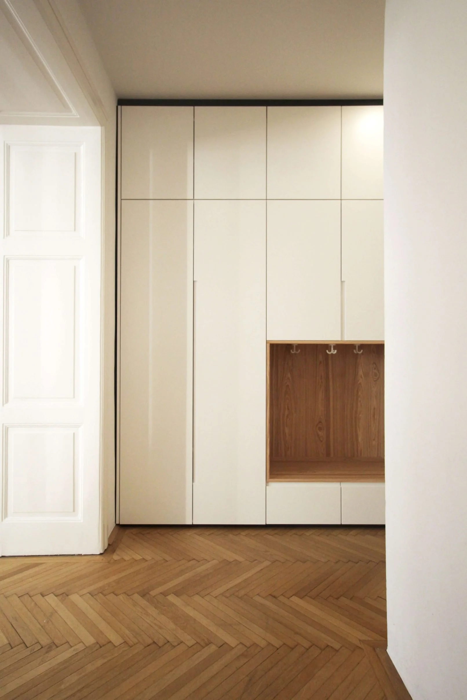 Modern white wardrobe with wooden interior hooks and storage, next to a white closed door and herringbone wooden floor.