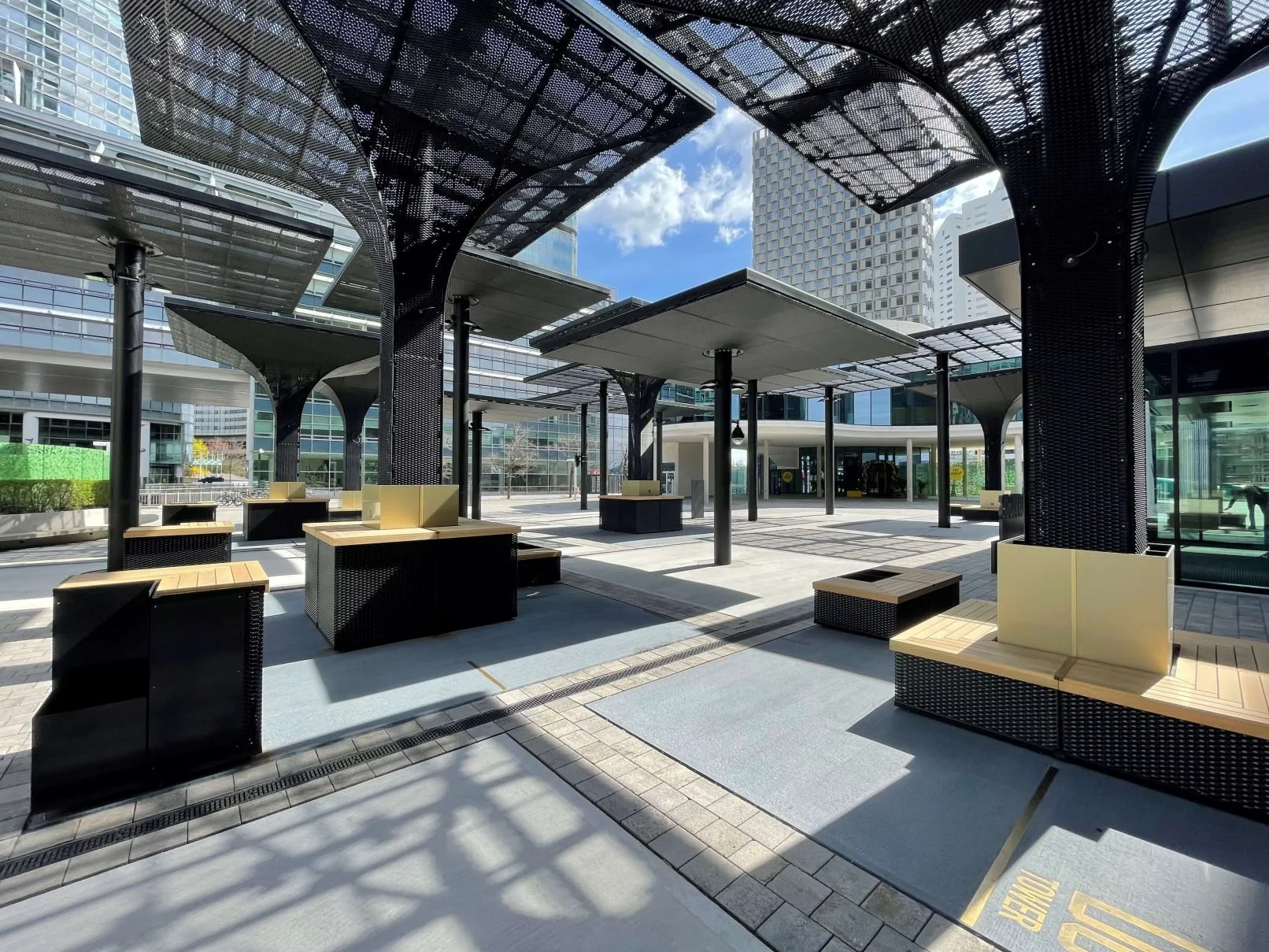 Empty outdoor terrace in an urban area with black benches, wooden seating, and modern black canopy structures, surrounded by tall glass office buildings.