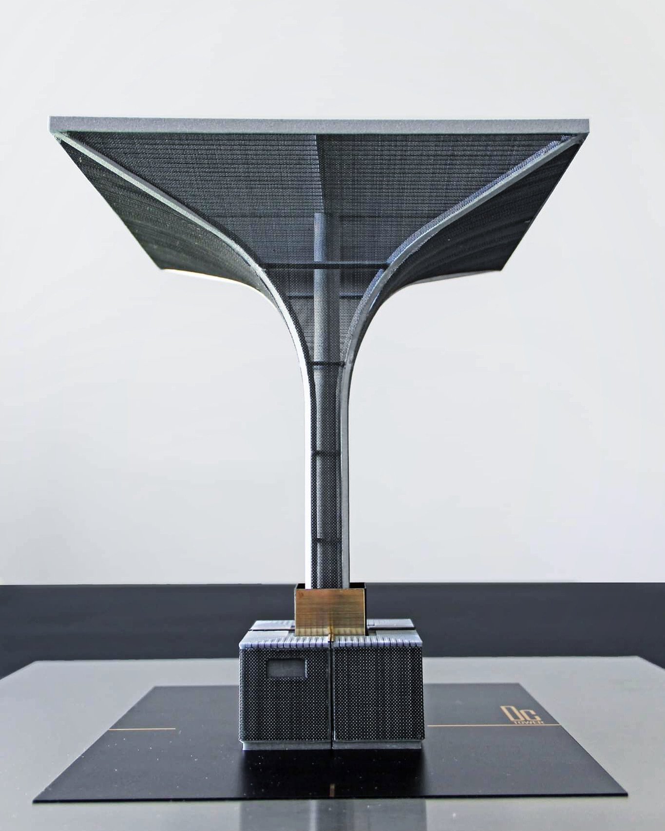 A modern architectural model with a central pole supporting a large, flat, umbrella-like structure made of black metal mesh, placed on a black base with a logo, against a plain white background.