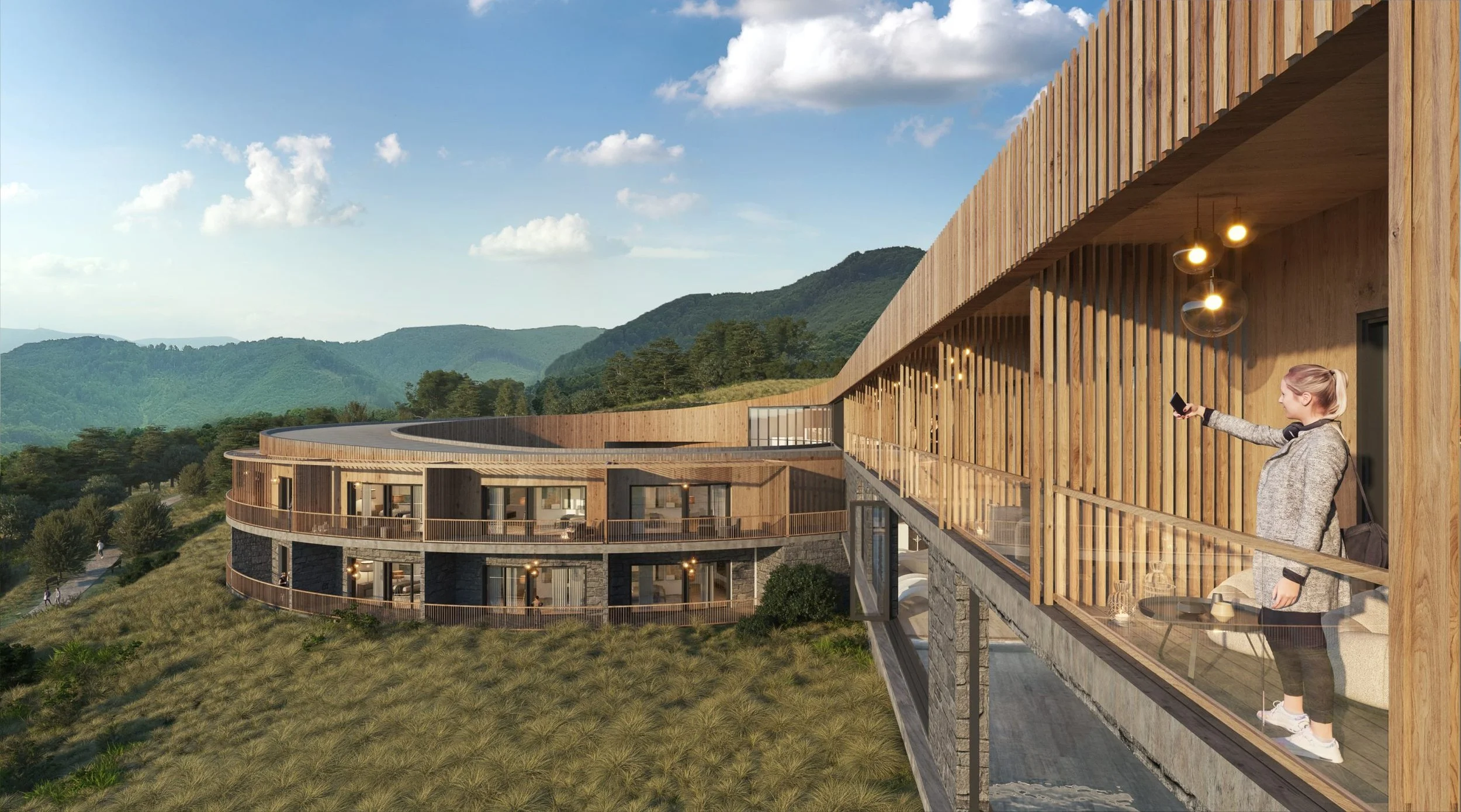 A woman standing on a wooden balcony of a modern, multi-story building with a mountain landscape in the background, taking a selfie with her phone.