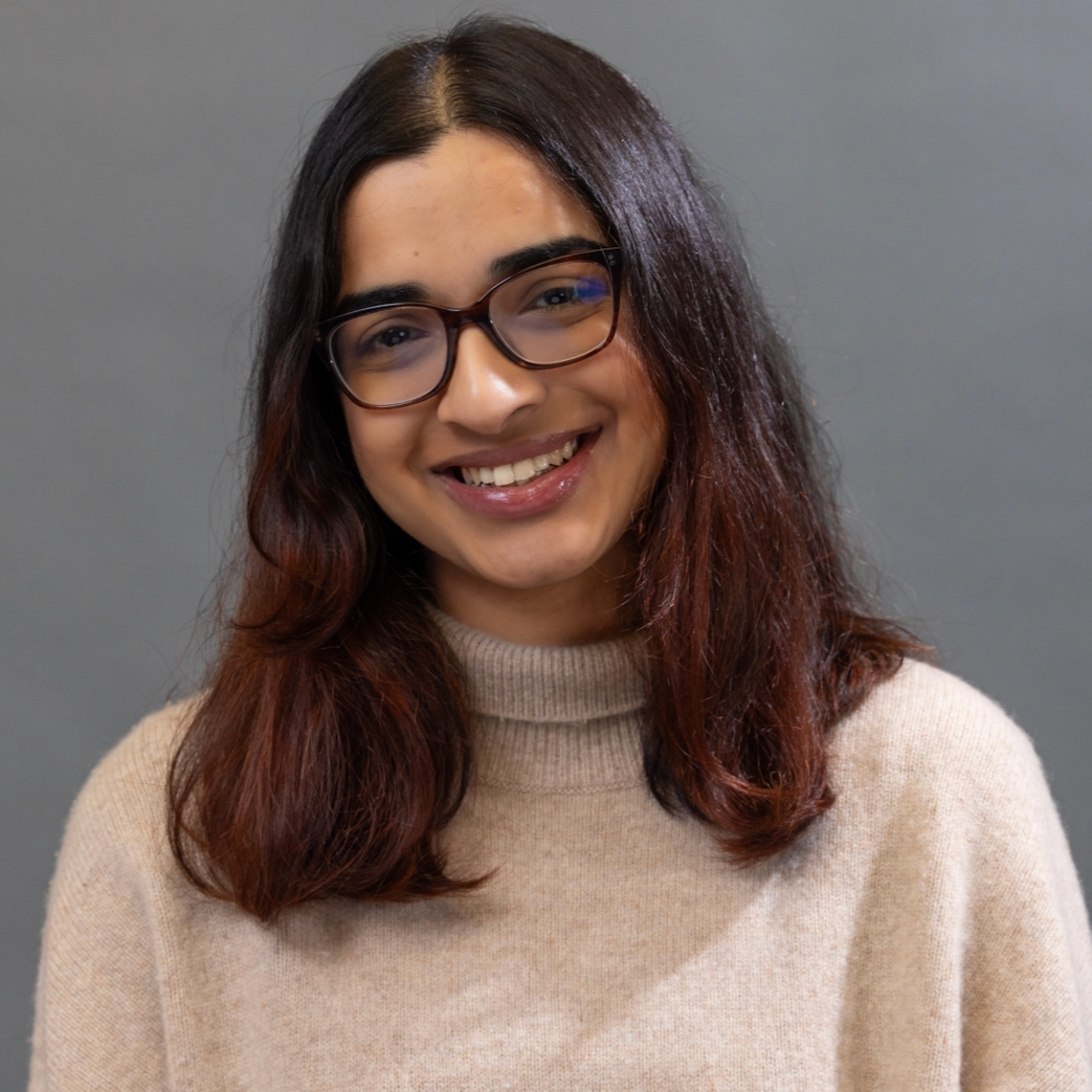 A young woman with shoulder-length dark hair and glasses, smiling in front of a plain gray background.