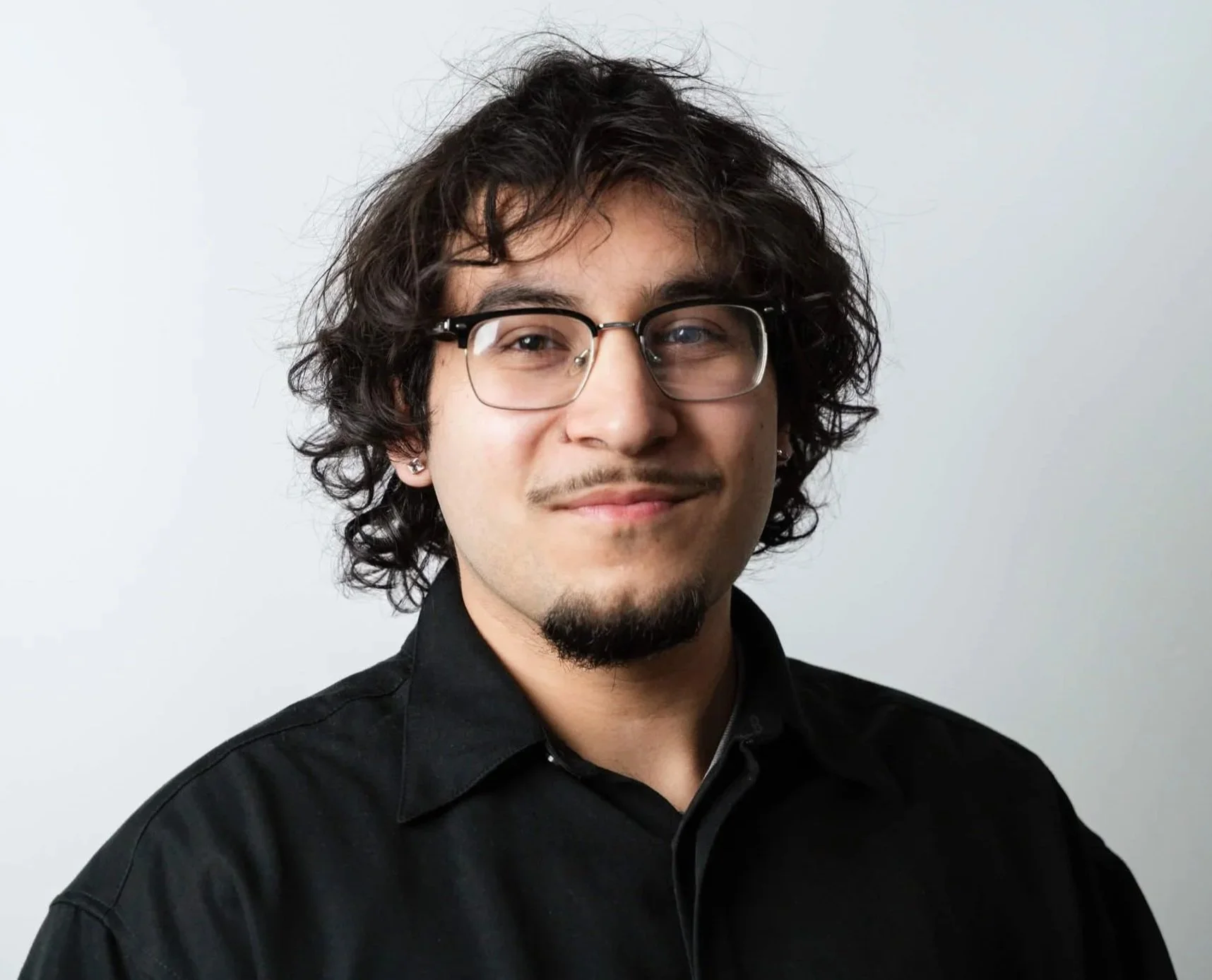 A young man with curly dark hair, glasses, light skin, wearing a black shirt, smiling slightly, against a plain light background.
