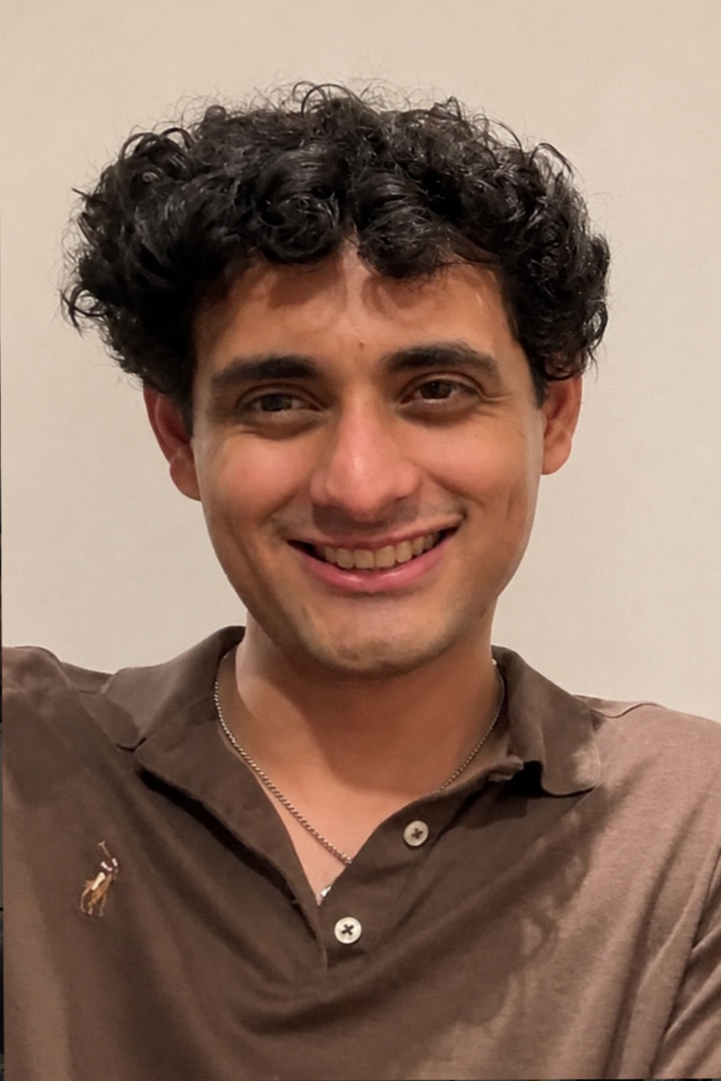 A young man with curly dark hair, smiling, wearing a brown polo shirt with a little giraffe emblem on the chest, and a silver necklace.