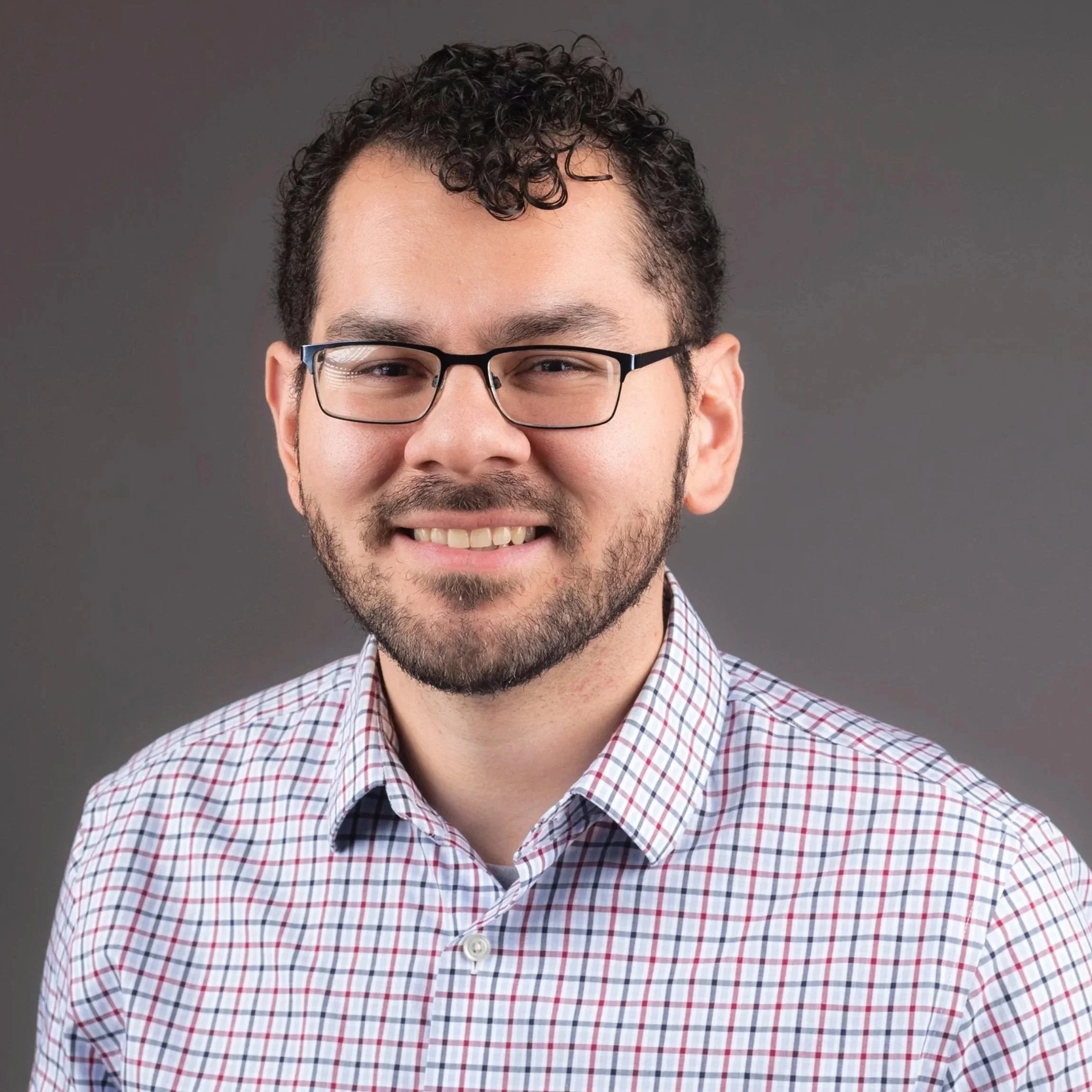 Close-up of a young man with curly dark hair, glasses, a trimmed beard, smiling, wearing a checkered shirt, against a plain gray background.