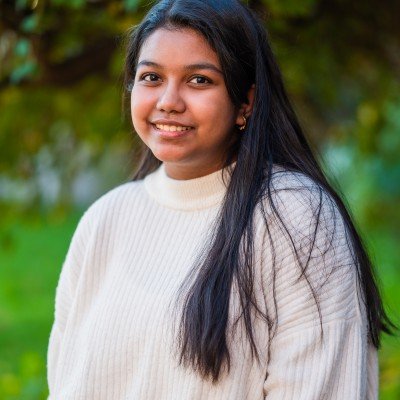 A young woman with long dark hair, wearing a cream-colored sweater, standing outdoors in front of green trees and grass, smiling at the camera.