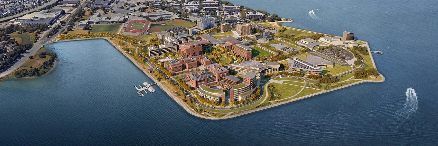 Aerial view of a university campus on a waterfront with buildings, sports fields, and green spaces surrounding the water.