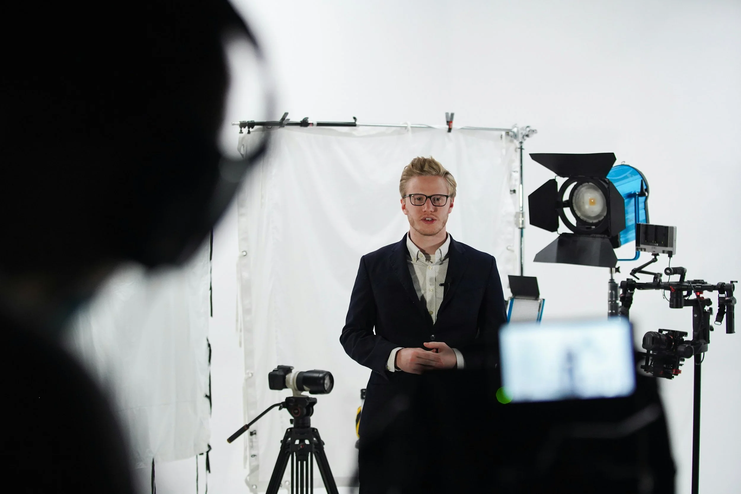 A man with glasses in a dark suit is standing in front of a white backdrop, being filmed with professional studio lighting and camera equipment.