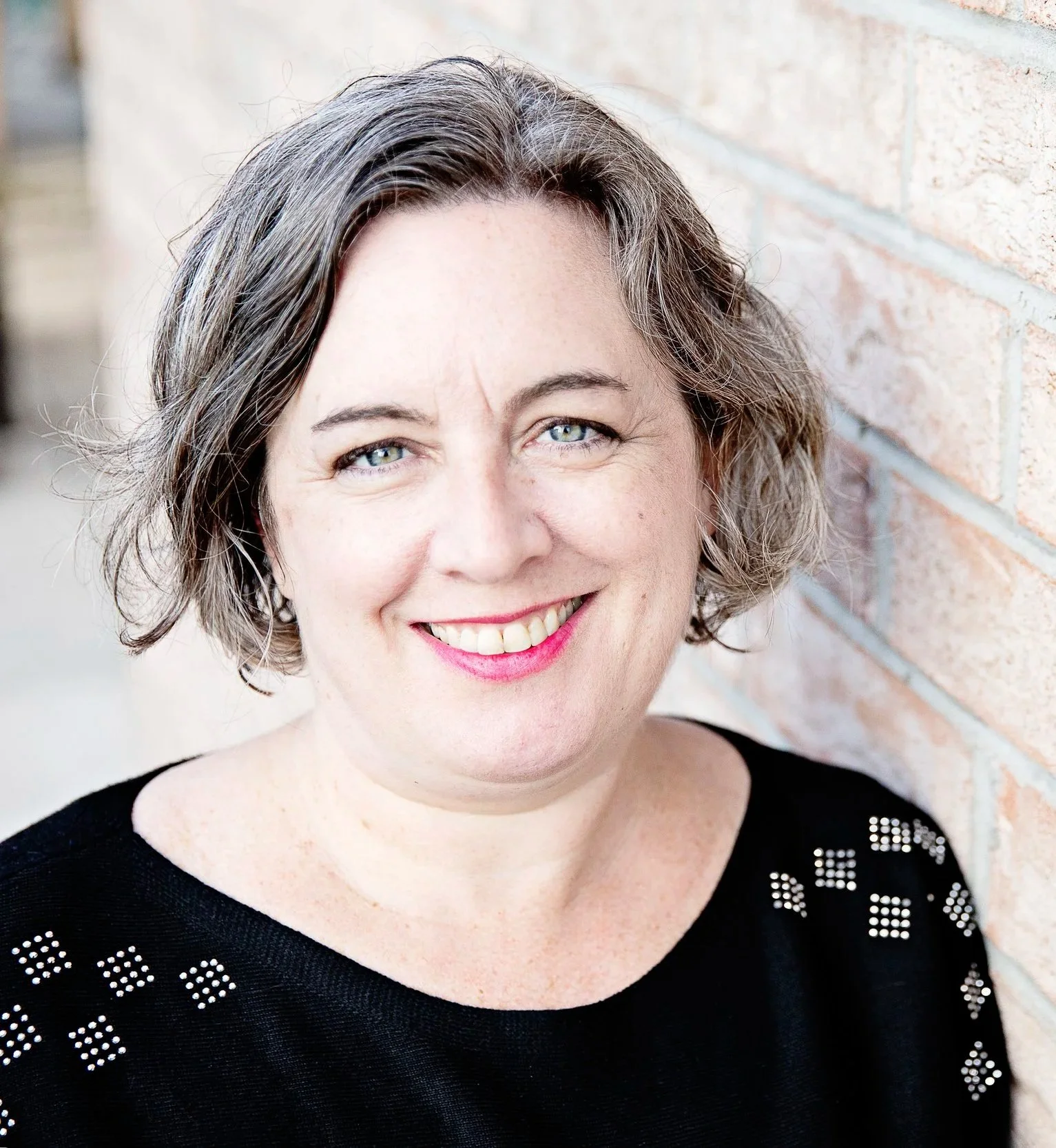Close-up of a smiling woman with curly short brown hair, wearing a black top with white dot patterns, sitting outdoors against a brick wall.